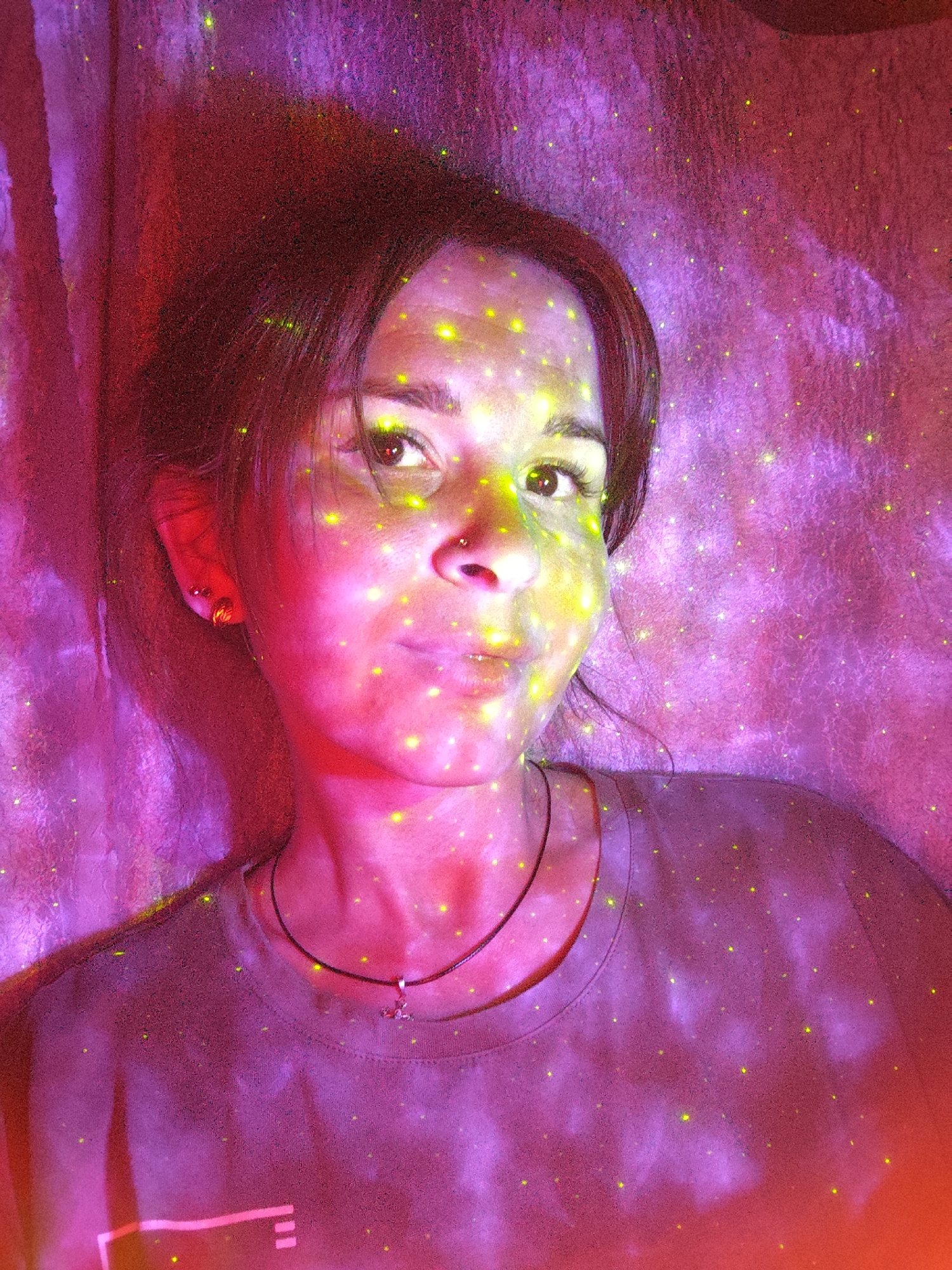 A female subject, an adult with dark hair, is depicted in a medium close-up, looking directly at the viewer. She has a small stud piercing on her left nostril and multiple stud earrings visible on her left ear. A dark cord necklace with a small metallic charm hangs around her neck, resting on a dark-colored, round-neck t-shirt. Her face and upper torso are illuminated by a combination of strong red, pink, and purple light, with numerous small, bright yellow-green dots projected across her skin, hair, clothing, and the background. The background consists of a textured surface, possibly a fabric or wall, exhibiting folds and variations in illumination, primarily in shades of purple and red, also covered by the yellow-green dots. The scene is located in the city of Фоки, Russia.