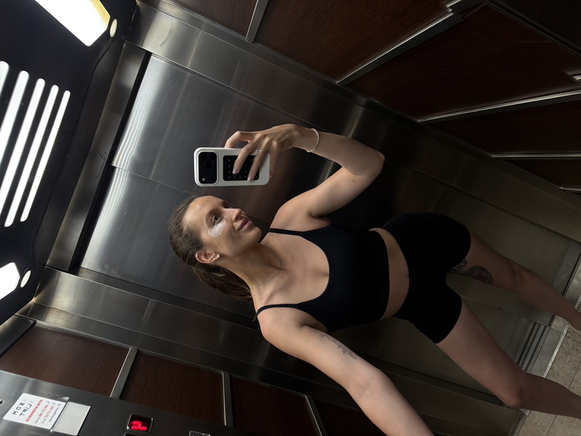 A woman is taking a selfie in an elevator. She is wearing a black sports bra and black shorts. She has under-eye patches on and a thin necklace. Her hair is pulled back in a ponytail. The elevator has wood-paneled walls and metal accents. There is a red illuminated button on the left side of the elevator. The woman's left arm has a tattoo. She is holding a white phone in her right hand. The scene appears to be inside an elevator in Budva, Montenegro.