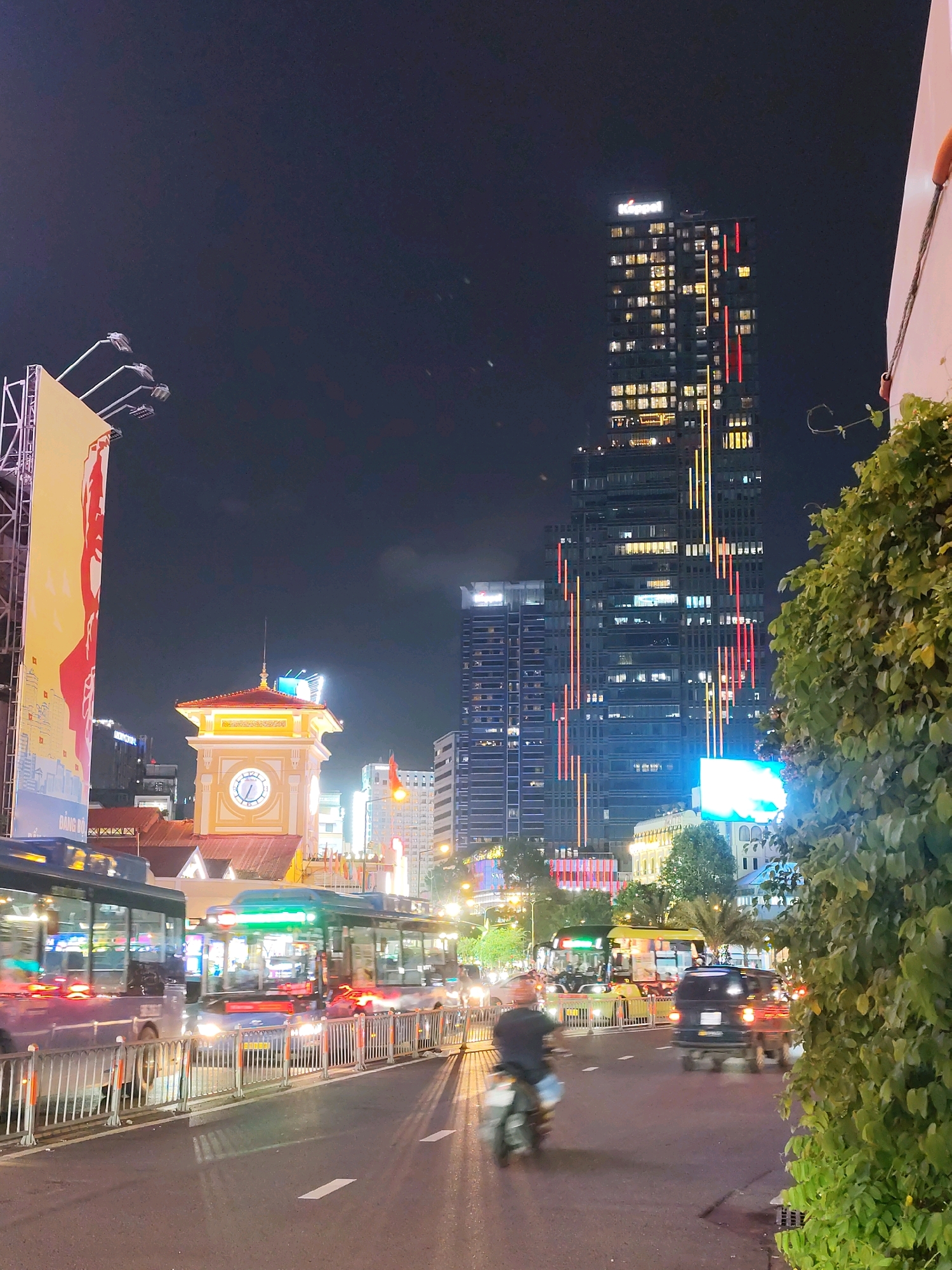 This image depicts a nighttime street scene in Ho Chi Minh City, Vietnam, specifically in front of the Saigon Central Post Office, also known as the Bưu điện Trung tâm Sài Gòn. A prominent clock tower, characteristic of the Post Office, is visible with its yellow facade and illuminated clock face. To the left, a large, brightly lit billboard stands, featuring a stylized red graphic.
The street is busy with traffic. Two blue buses are in the foreground, their headlights and interior lights creating streaks of light due to motion blur. Several other vehicles, including a dark minivan and motorcycles, are also present. A single motorcycle rider is moving away from the camera.
In the background, a modern skyscraper with many illuminated windows dominates the skyline. Vertical lines of red and yellow lights are visible on its facade, and a sign at the top reads "Keppel." Other buildings, some with illuminated signage and architectural features, are interspersed throughout the cityscape. Lush green foliage from a nearby tree is visible in the right foreground, partially obscuring the view of the buildings and the street. The sky is dark, indicating it is nighttime, and streetlights cast a warm glow on the wet pavement.