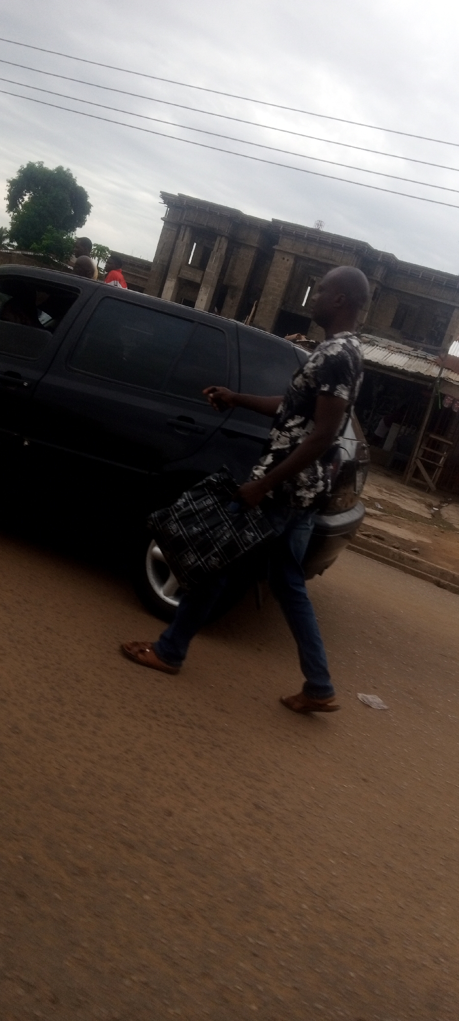 A man in a floral shirt and jeans is walking on a street in Sabon Tasha, Nigeria, next to a black car. He is carrying a black and white checkered bag. The street is paved and there are buildings in the background, including one that appears to be under construction.