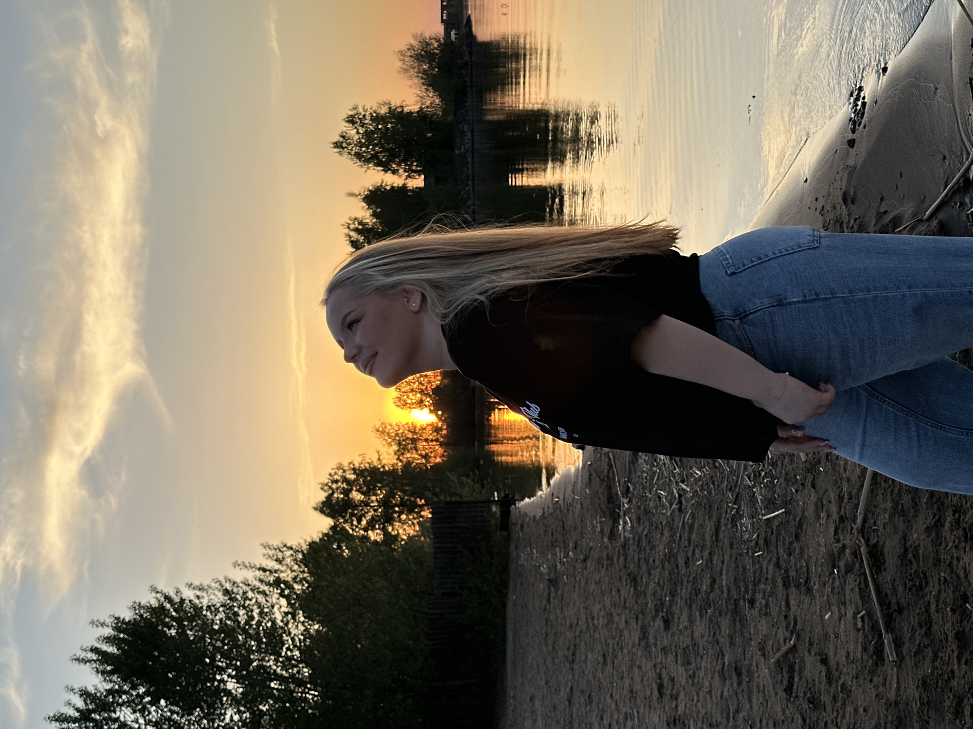 A young woman with blonde hair is standing on a sandy beach next to a body of water, likely a river or lake, at sunset. She is wearing a black shirt and blue jeans. The setting sun is visible through the trees on the opposite shore, with its reflection visible in the water. The location is likely near Chaikovsky, Russia, given the prompt.