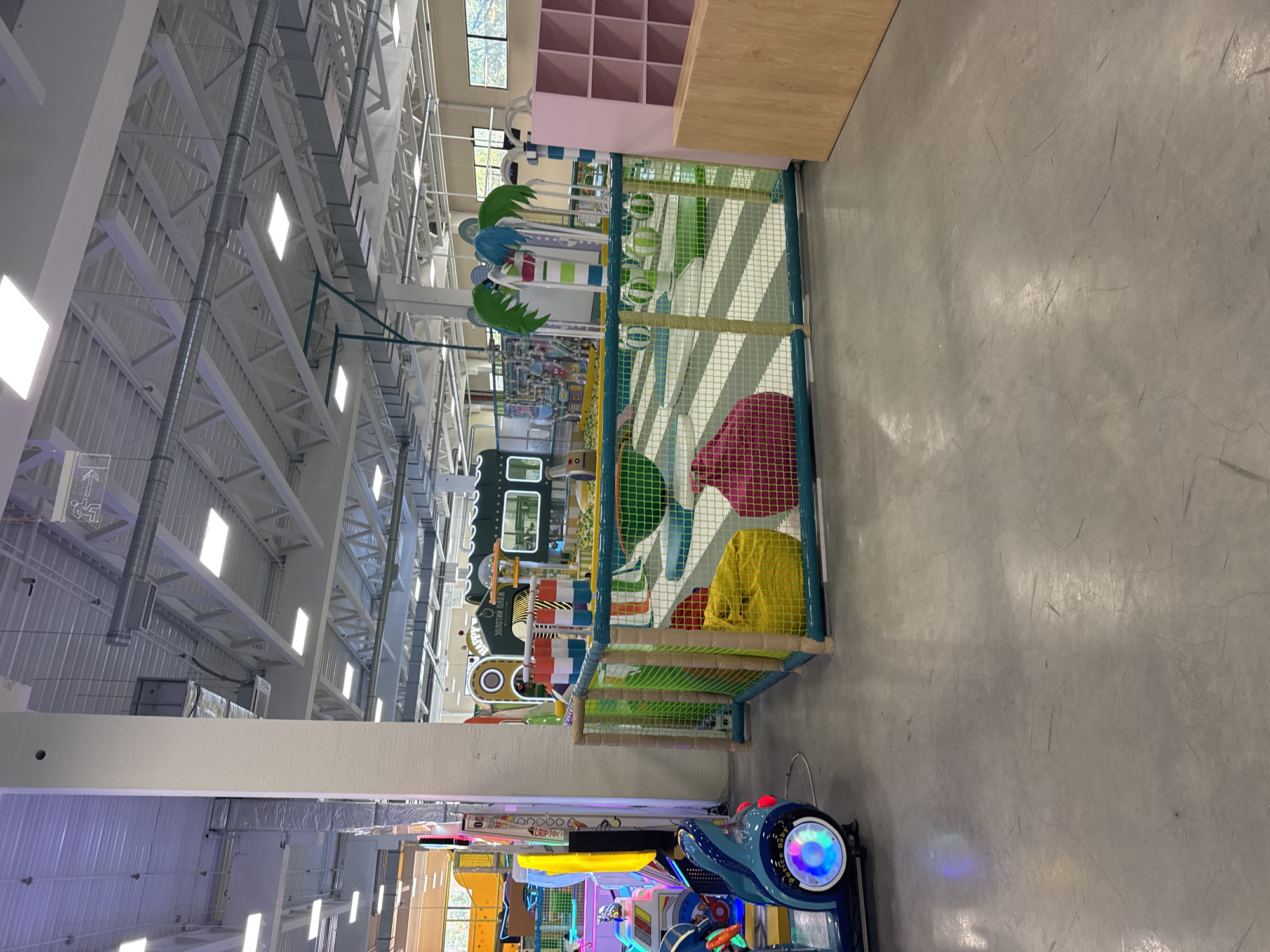The image depicts an indoor children's play area, possibly at a mall or entertainment center, located in Orlivshchyna, Ukraine. The space is brightly lit with overhead fluorescent lights and features a variety of attractions. In the foreground, a blue and black rideable toy, resembling a car with flashing lights and the text "Police Baby" visible, is parked on a gray concrete floor. Adjacent to this is a soft play area enclosed by a green net fence with tan posts. Inside this enclosure, a bright yellow beanbag chair and a pink beanbag chair are visible, along with several green and white striped balls scattered on a white and green patterned soft surface. In the background, there are more play structures, including a green train-like building with windows and the text "HAPPY" and "ЗОЛОТИЙ ПЛЯЖ" (Golden Beach) visible. Other colorful play elements and a large pink shelving unit are also present. The ceiling is a complex network of white beams, ducts, and lighting fixtures. The overall environment suggests a recreational facility designed for children.