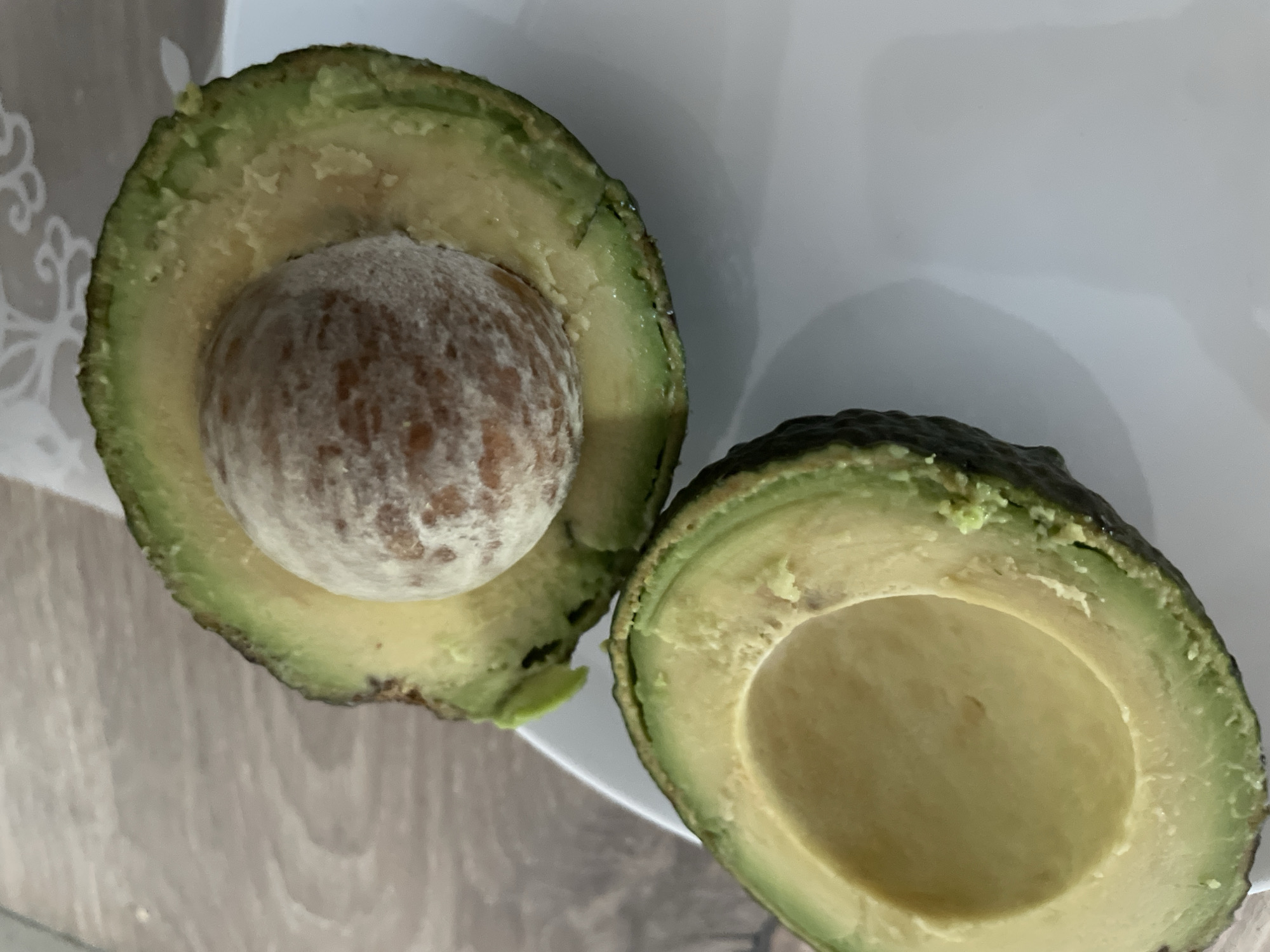 The image shows a close-up, overhead view of a halved avocado on a white plate. The avocado is cut lengthwise, revealing its green flesh and a large, round pit in one half. The other half is empty, showing the cavity where the pit was. The avocado sits on a white plate, which is placed on a wooden surface. The background shows a light-colored fabric with a patterned texture. The image does not contain any information or context that would allow for the determination of its location in Brovary, Ukraine.