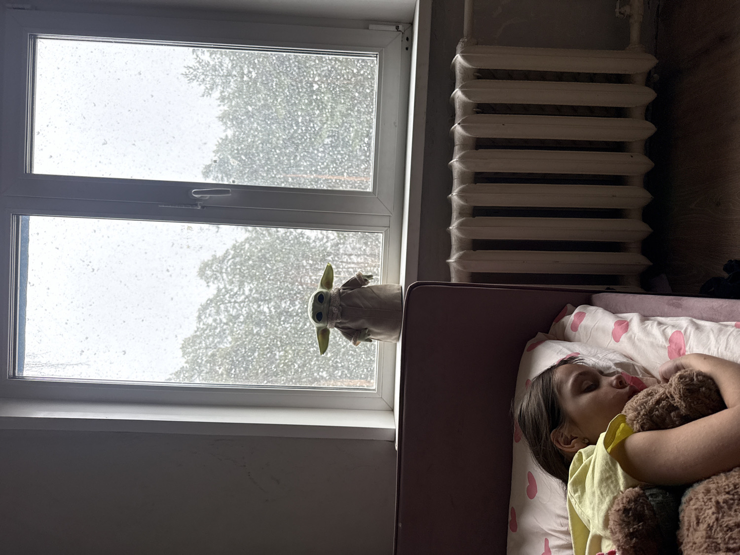 The image displays an indoor scene, likely a bedroom, with a young girl sleeping peacefully in a bed. The girl is lying on her side, wearing a yellow top, and embracing a brown teddy bear. Her head rests on a white pillow patterned with pink hearts, and the bed features a dark pink headboard.
To the right of the bed, a two-pane window reveals an outdoor view. The window glass is speckled with droplets, indicating rain or light snow, and blurred green tree foliage is visible beyond. A "Baby Yoda" (Grogu) plush toy sits on the window sill, facing outwards. Adjacent to the window, a white, ribbed radiator is mounted on the wall.
The room is dimly lit by natural light filtering through the window, suggesting an overcast day or early morning. The weather outside is characterized by precipitation. The overall atmosphere is quiet and calm, with the girl resting comfortably indoors. No text is visible in the image. The setting is consistent with a residential interior in Votkinsk, Russia.
