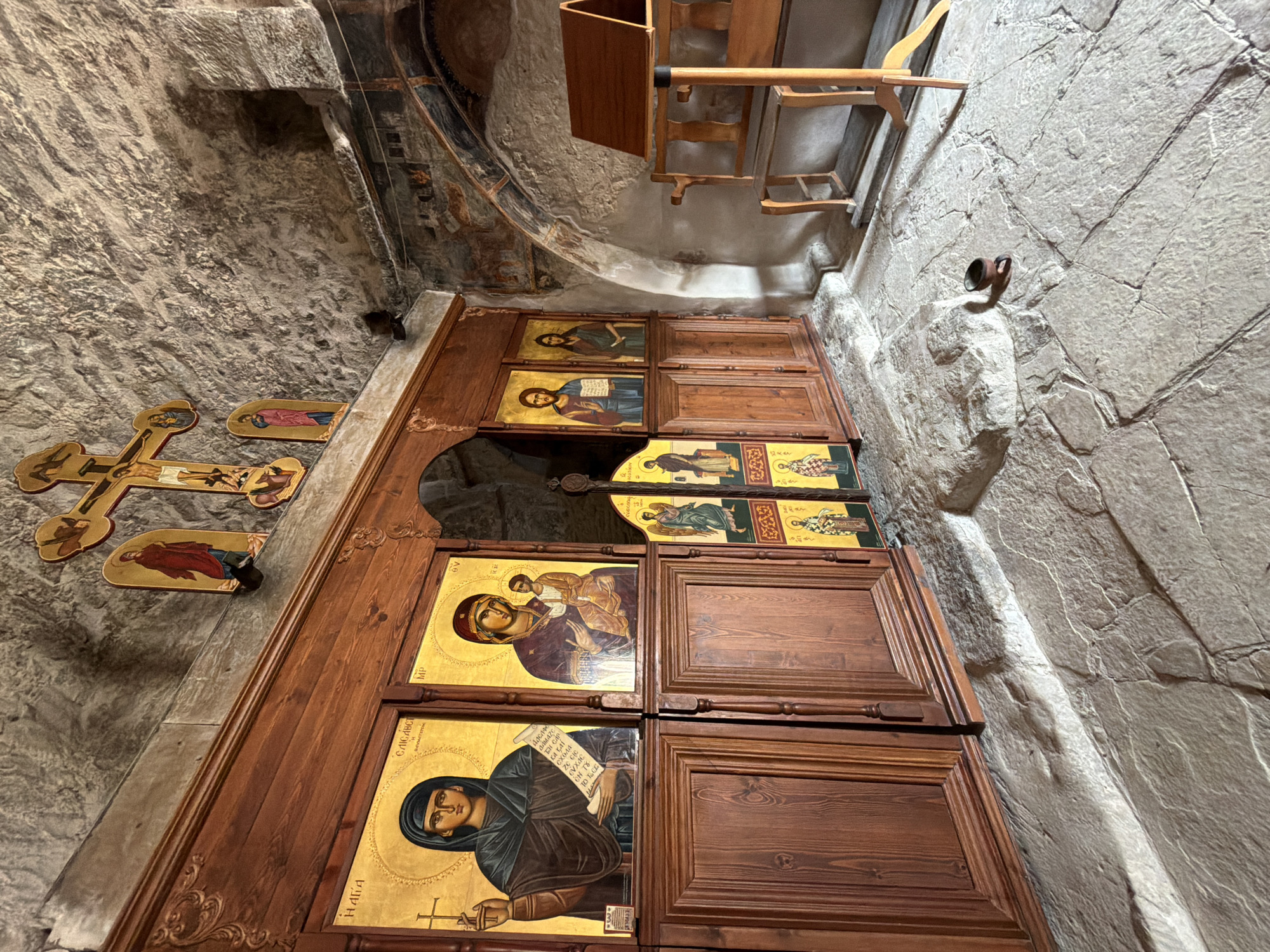 The image depicts the interior of a Byzantine-style church or chapel in Agios Therapon, Cyprus. The focal point is an iconostasis, a wall of icons, made of dark wood. Several framed religious icons are mounted on the iconostasis, featuring figures like the Virgin Mary, Jesus Christ, and other saints. The icons are rendered in a traditional, flat style with gold backgrounds. In the upper part of the scene, a large, ornate cross is mounted on the stone wall, adorned with smaller icons of religious figures. The walls of the structure are made of rough, light-colored stone, suggesting an ancient or traditional architectural style. To the right, a wooden lectern or stand is positioned, designed to hold books or other religious texts. The floor is paved with irregular, large stone slabs, and a small, dark cup rests on the floor in the foreground. The lighting is subdued, casting shadows that highlight the texture of the stone walls and the raised surfaces of the icons. The overall atmosphere is one of solemnity and religious reverence.