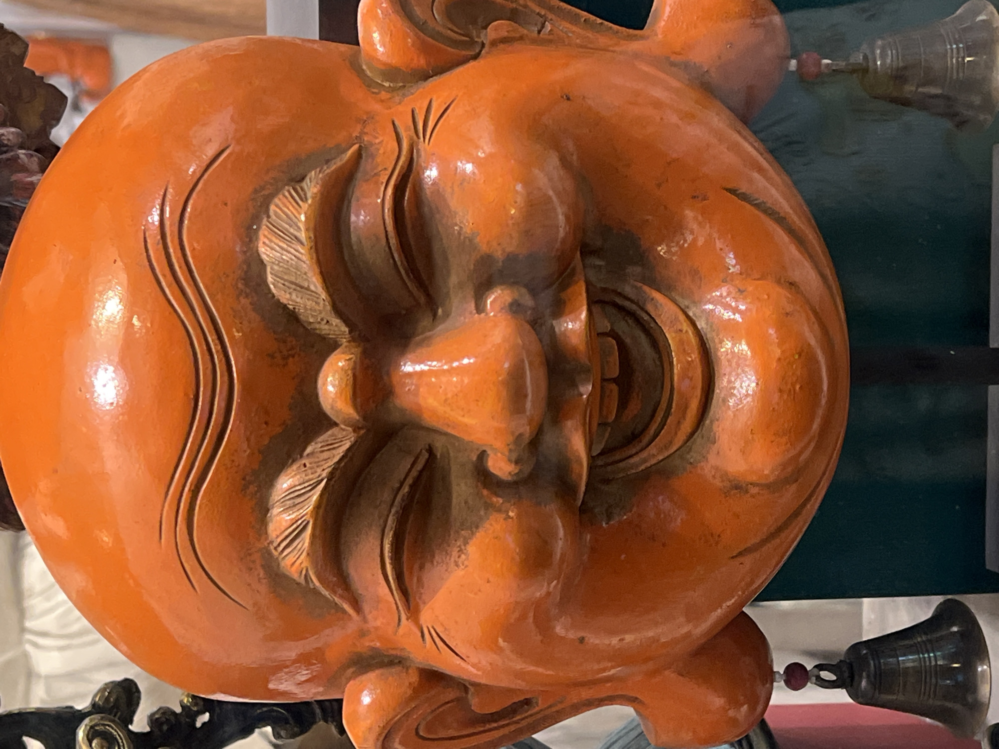 The image presents a close-up view of an orange-colored, carved wooden sculpture of a face. The face appears jovial, with exaggerated features such as prominent cheekbones, a wide nose, and a partially open mouth revealing teeth. The carving includes stylized details around the eyes and cheeks, suggesting traditional artistry. The sculpture is glossy, reflecting light and highlighting its textures. In the background, blurred elements include metallic-looking objects and a dark green surface. Based on the "Ubud District, Indonesia" information, this is likely a cultural artifact or decorative piece found in that region, known for its wood carving traditions.