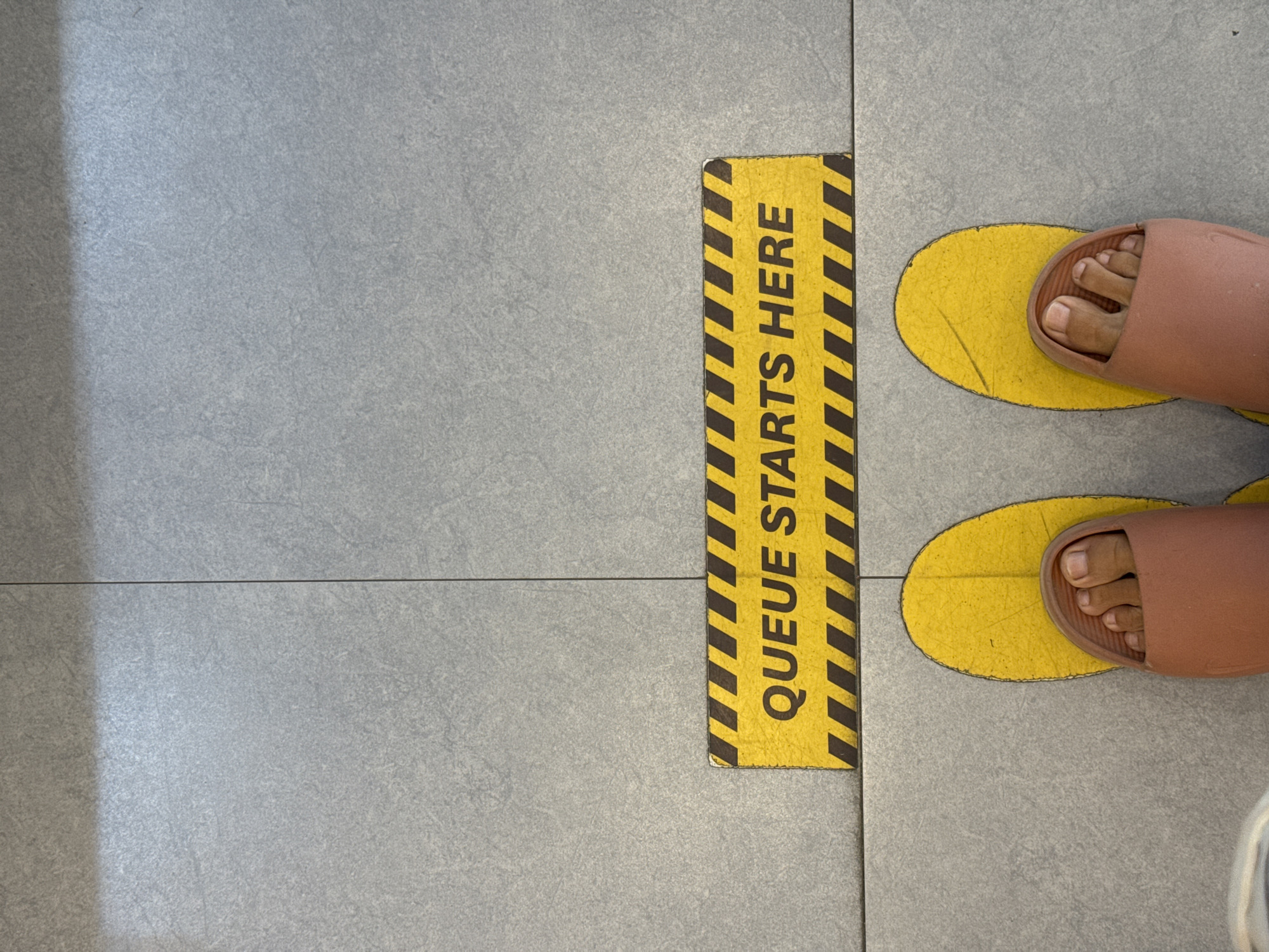 The image shows a person standing in a queue, indicated by a yellow and black striped sign that reads "QUEUE STARTS HERE" on a tiled floor. The person is wearing light pink sandals and standing on yellow oval foot markers. The location is in Kuta Selatan, Indonesia.