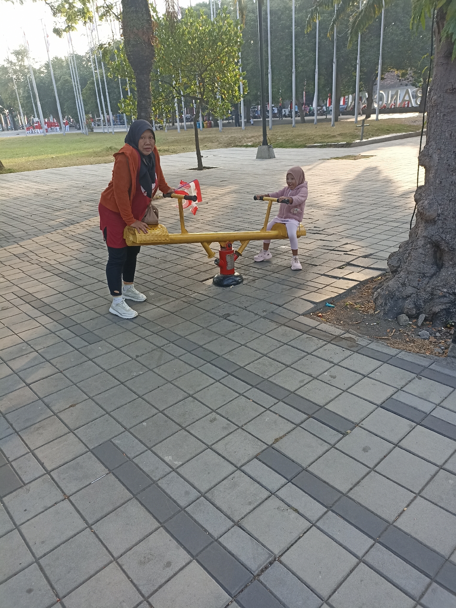 The image depicts an outdoor scene with a woman and a young child on a yellow seesaw. The woman, wearing an orange jacket and a headscarf, is standing on the ground next to one end of the seesaw. The child, wearing a pink outfit and a headscarf, is seated on the other end of the seesaw, holding onto the handles. The seesaw is positioned on a paved area with square gray tiles. In the background, there are trees, flagpoles, and some people walking in a park-like setting. The overall lighting suggests it is daytime, likely in the morning or late afternoon due to the warm tones and shadows. The scene appears to be within a public space in Kelurahan Kepatihan, Indonesia, consistent with a city park or recreational area.