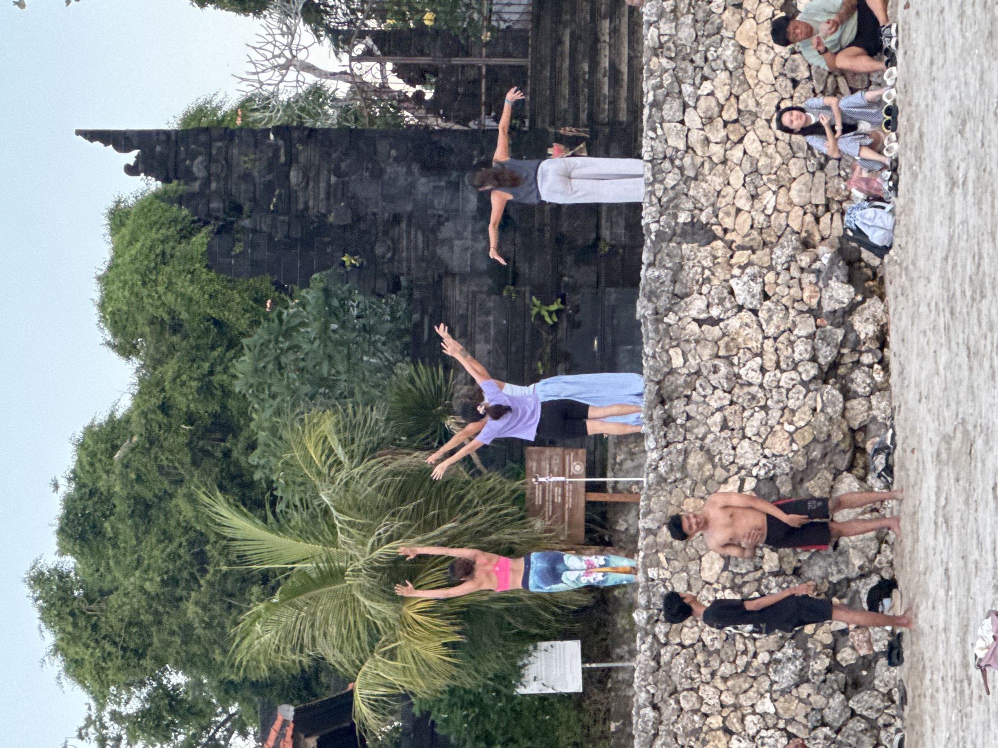 In Kuta Selatan, Indonesia, a group of people are gathered on a sandy beach in front of a stone wall and a weathered stone structure. Three women are engaged in what appears to be a yoga or meditation session on top of the wall, with their arms outstretched. Behind them, a dense canopy of trees and a palm frond are visible, framing the scene. To the right of the stone structure, there are steps leading up to an entrance. On the beach, two young men are conversing, one shirtless and the other wearing a black t-shirt and shorts. Further to the right, a seated woman and man are also present on the sand, with several pairs of footwear and bags nearby. The overall setting suggests a relaxed, outdoor activity taking place in a coastal environment.