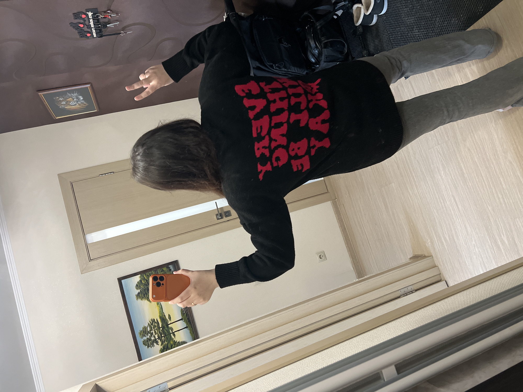A person stands in front of a mirror, taking a selfie with an orange-cased smartphone. The individual is wearing a black sweater with red lettering on the back and grey pants. A black backpack is slung over one shoulder. The floor is light-colored wood. A painting hangs on the wall behind the person, and another smaller painting is on the wall above the door. The location is a room, potentially in a home in Perm, Russia.