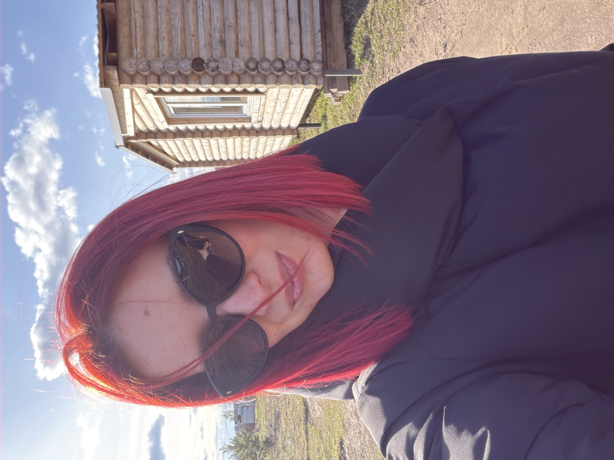 The image shows a person with red hair wearing sunglasses and a dark jacket. They are positioned in front of a wooden building, possibly a cabin or house, under a blue sky with clouds. The ground appears to be a mix of grass and dirt.
Based on the information that this image was taken in the city of Chaikovsky, Russia, the wooden building could be a traditional Russian structure often found in rural or suburban areas. The environment suggests an outdoor setting, possibly near a park, countryside, or outskirts of the city. There are no evident actions or interactions in this image.