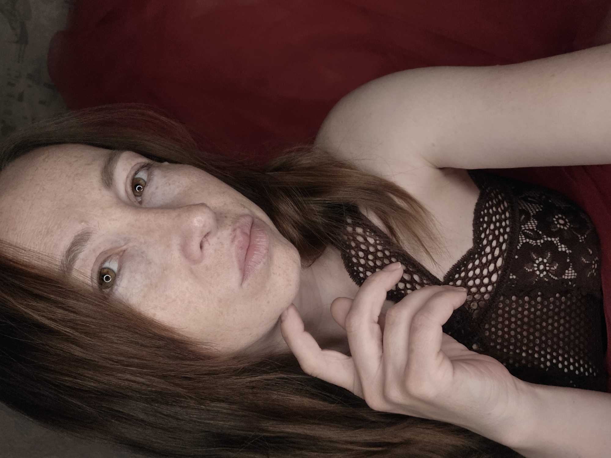 A woman with light skin and freckles is positioned against a red backdrop, possibly fabric. She is lying down, with her head tilted to the left. Her hair is brown, and she has brown eyes. She is wearing a dark lace top. Her right hand is near her face, with fingers touching her chin. The environment appears to be indoors. There is no indication of a city or location.