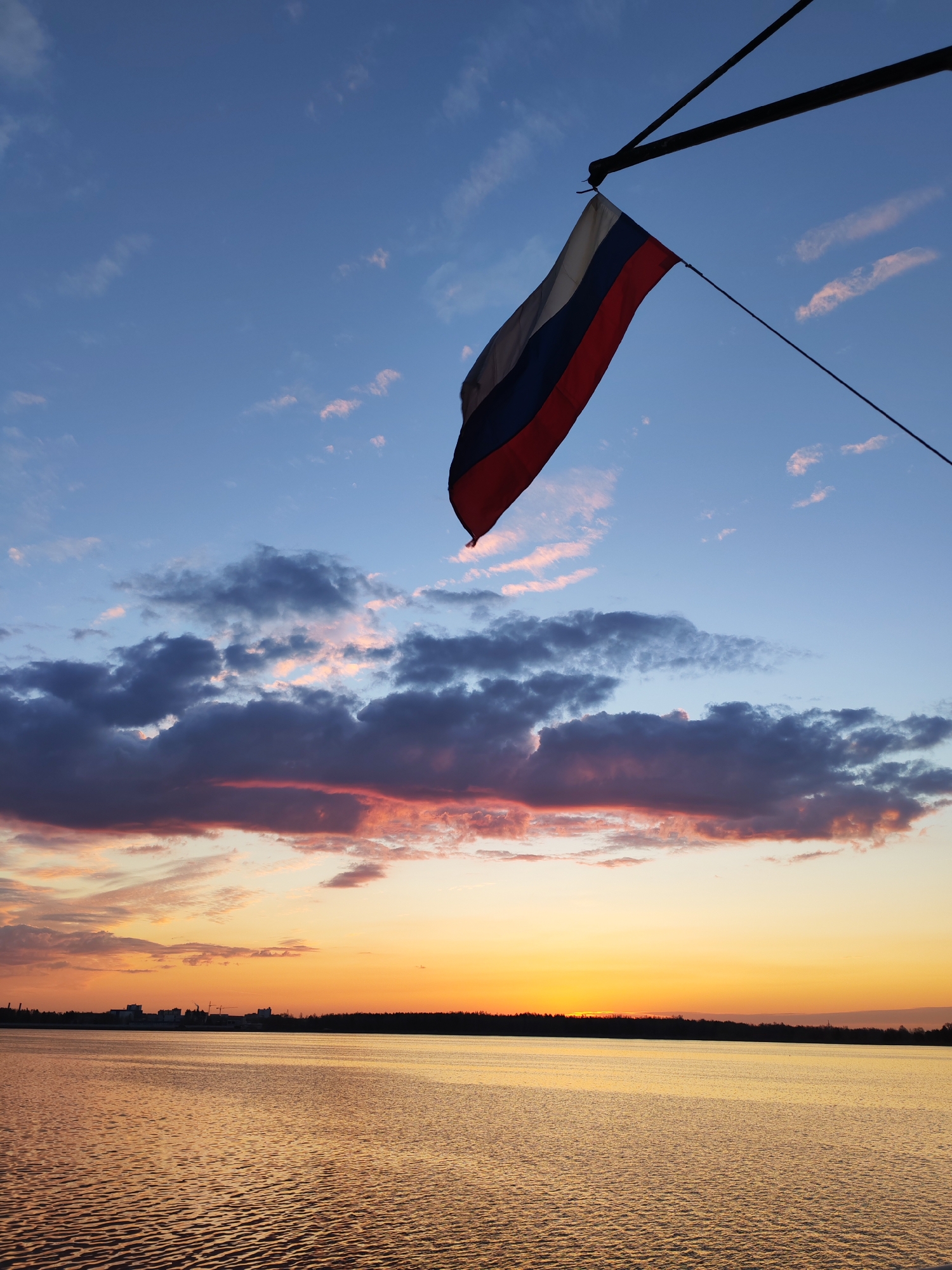 The image captures a Russian flag hanging from a pole against a colorful sunset sky. The sky transitions from blue at the top to orange and yellow near the horizon, with clouds scattered throughout. The bottom of the image features a body of water reflecting the colors of the sunset, and a dark treeline and some buildings are visible on the far shore. The location is Unknown, Unknown.