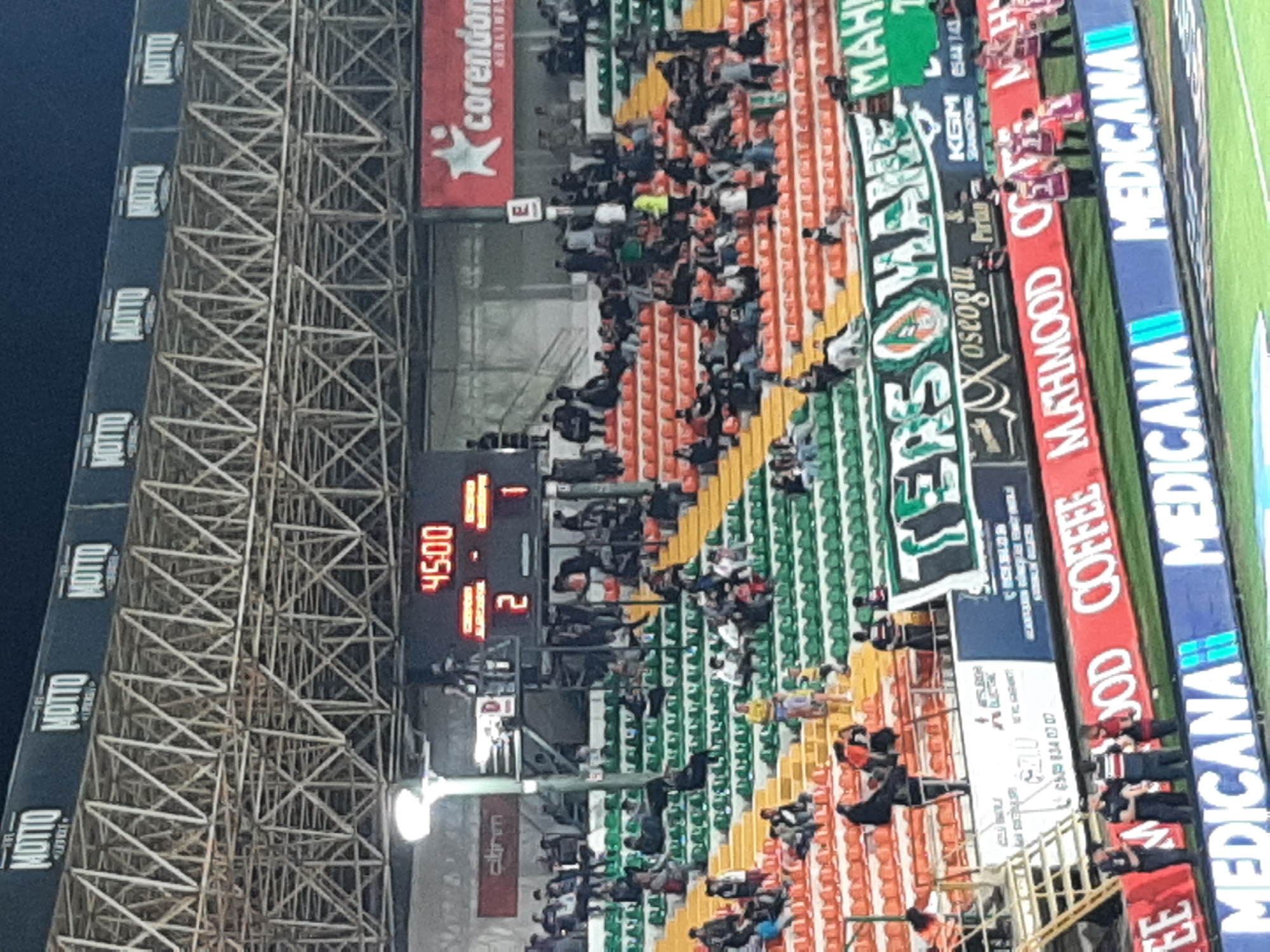 The image depicts a stadium scene, likely during a soccer/football match. The score on the scoreboard reads 1-2, with the time at 45:00, signaling the end of the first half. Spectators are seated in orange and green stadium seating, with a mix of activity and rest. Banners displaying "TERSA" and "MANE" are visible, along with advertising boards that feature "COFFEE MAHMOOD" and "MEDICANA". The stadium structure is apparent, with scaffolding supporting lighting and advertising displays including the word "MOTTO". Based on the presence of the "Korendon" airline advertisement, it is possible that this image was taken at the Alanya Oba Stadium in Alanya, Turkey.