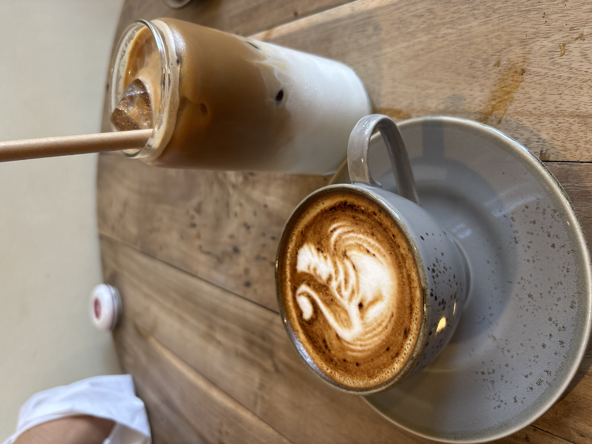 Two coffee drinks sit on a wooden table. One is in a glass, filled with ice and a layered coffee drink with a straw, lying on its side. The other is a latte in a speckled grey mug with a latte art design, sitting on a matching saucer. The background suggests an indoor setting, possibly a cafe. A white object with a red center is visible in the lower left corner. Based on the image, it is not possible to determine the exact location or confirm it is within Kuta Selatan, Indonesia.