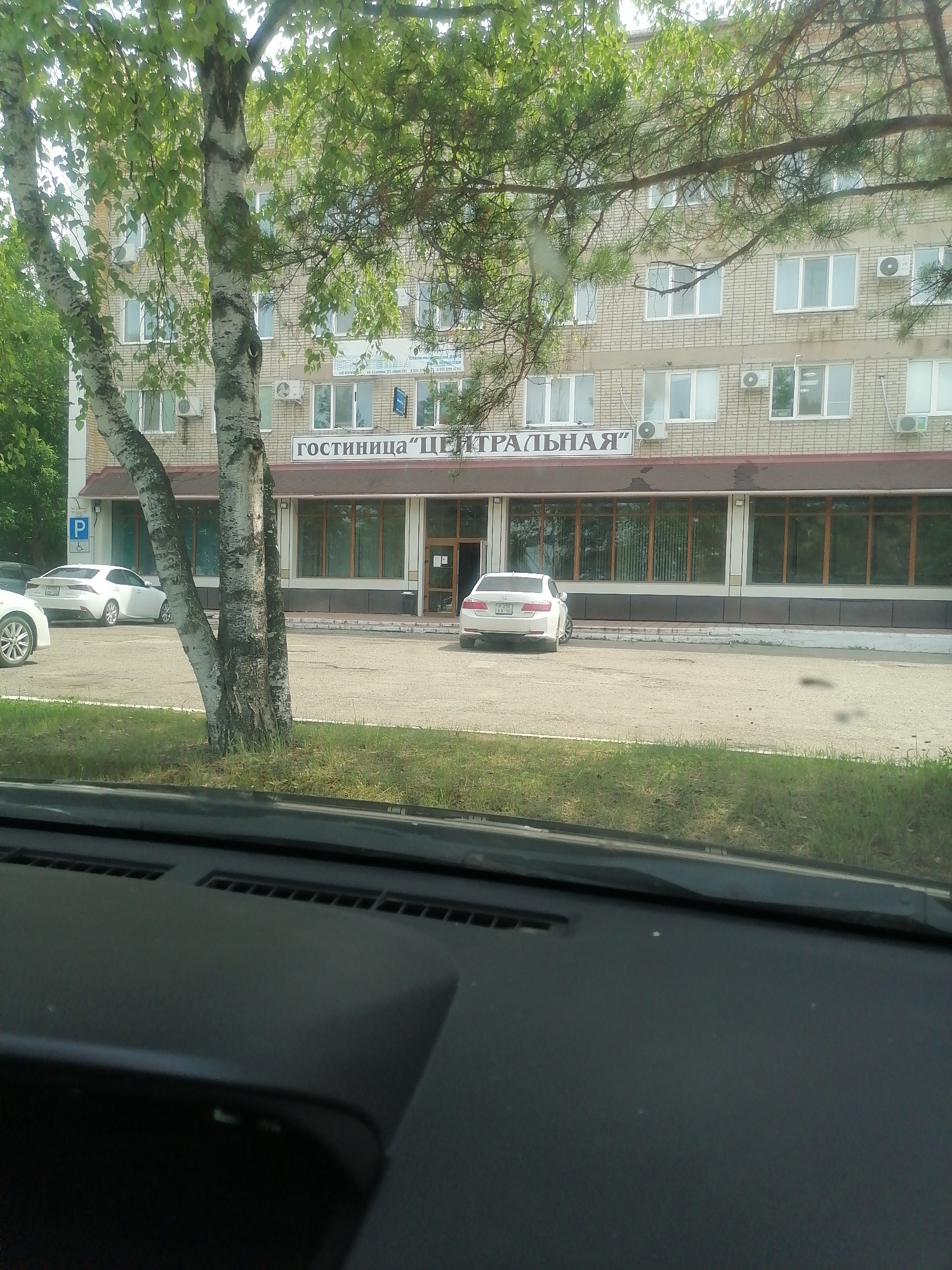 The image shows a white sedan parked in front of a brick building with the sign "ГОСТИНИЦА "ЦЕНТРАЛЬНАЯ"" (Hotel "Centralnaya") above the ground floor windows. The building appears to be a hotel, with several windows visible on multiple floors. There are also trees in the foreground, partially obscuring the view of the building. A parking sign with a "P" symbol is visible to the left of the entrance. The scene appears to be an outdoor setting, likely a street or parking lot, with grass and pavement visible. The photograph seems to have been taken from inside a vehicle, as indicated by the presence of a dashboard in the foreground. This location is identified as the city of Lesozavodsk, Russia.