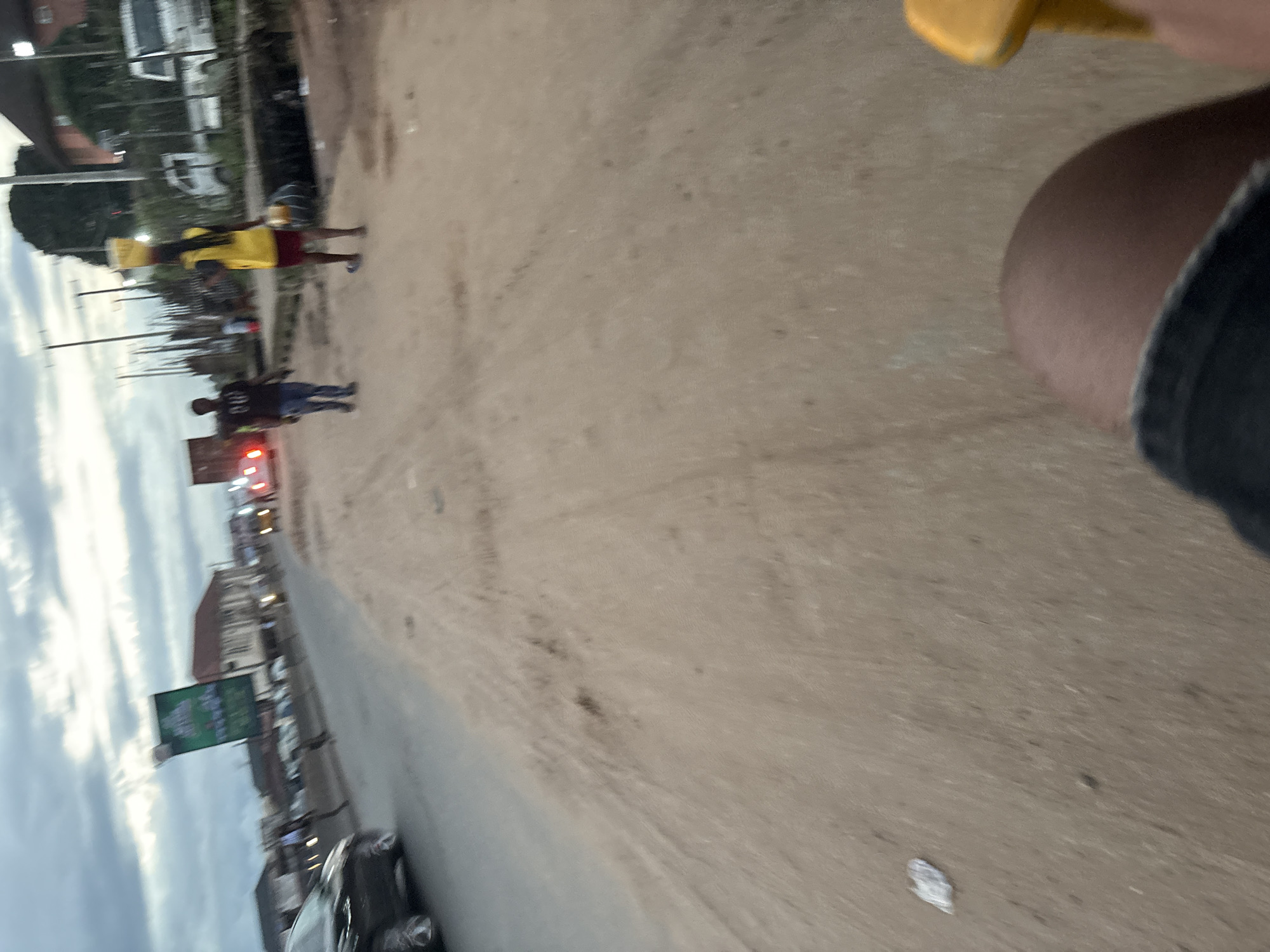 The image shows a dusty, unpaved road with sparse vegetation and buildings in the background. The sky is overcast. In the foreground, a person's knee and thigh are visible, suggesting the photo was taken from a low vantage point, possibly from a moving vehicle or bicycle. In the middle ground, a person in a yellow top and red shorts carries a large container on their head, walking away from the camera. Another person in a dark shirt and jeans is walking towards the camera. Several vehicles are visible on the road, including cars and trucks. A large billboard advertising a product is also present on the left side of the road. The scene appears to be a busy commercial or residential area in Onitsha, Nigeria, characterized by its unpaved roads and the presence of street vendors.