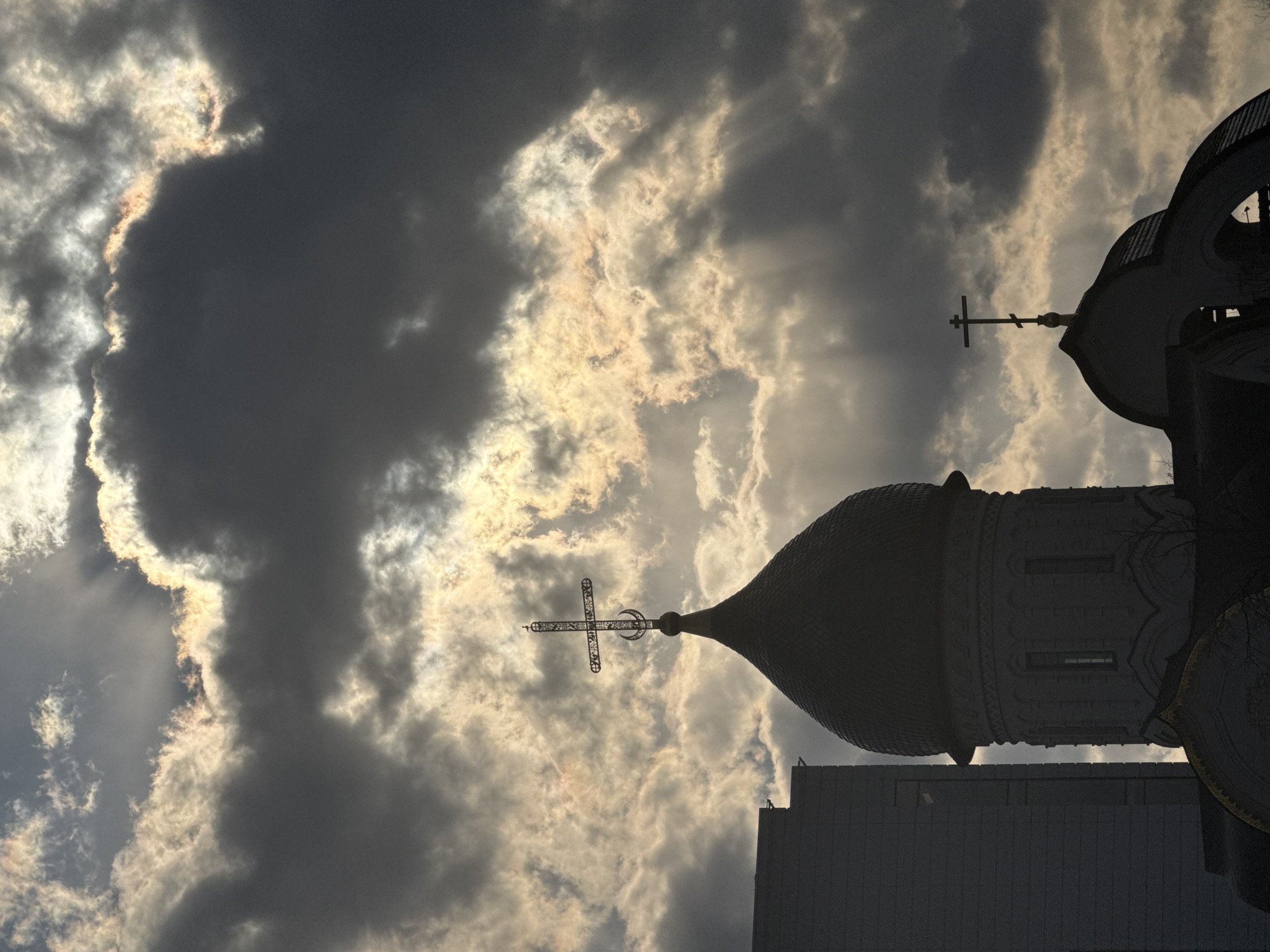 The image, taken from a low angle, features a silhouette of an Orthodox church against a dramatic sky filled with dark, textured clouds and visible sun rays. The church is a portion of the Moscow Cathedral Mosque, located in Moscow, Russia. Two domes of the Mosque are visible, each topped with a cross. The background is filled with a cloudy sky with dramatic sun rays.