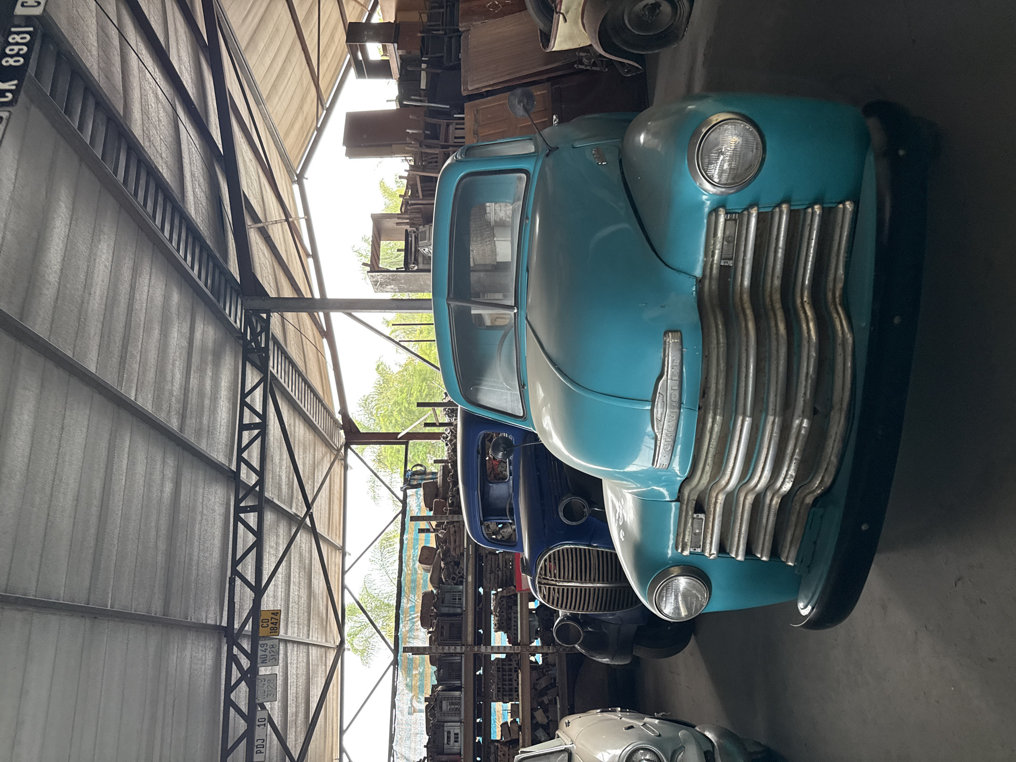 The image shows a warehouse or garage setting in Kraaifontein, South Africa, housing a collection of vintage vehicles. Prominently featured is a light blue Chevrolet pickup truck, with its front grill and headlights clearly visible. Behind the blue truck, a dark blue truck of similar vintage is partially visible. In the background, shelves are filled with various automotive parts and other items. The structure of the building includes metal support beams and a corrugated metal roof. Several vehicle registration plates are affixed to a support beam.