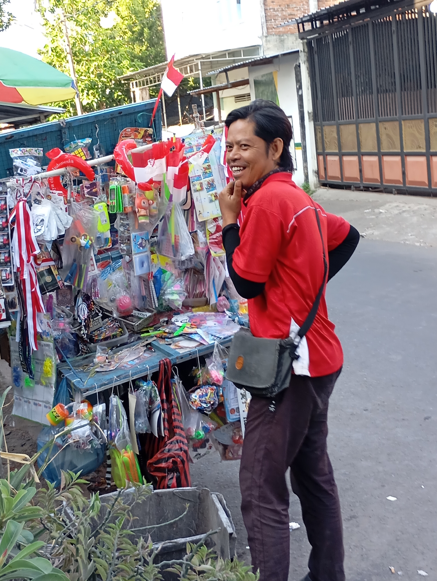 A man wearing a red shirt with black long sleeves and dark brown pants stands near a stall selling various items, including Indonesian flags. He is looking towards the left of the frame, with his right hand touching his chin and a smile on his face. The stall is filled with packaged goods, toys, and small flags, and is positioned on a paved street in Mataram, Indonesia. In the background, there is a building with a brick facade and a metal gate. The scene appears to be outdoors, with sunlight illuminating the area.