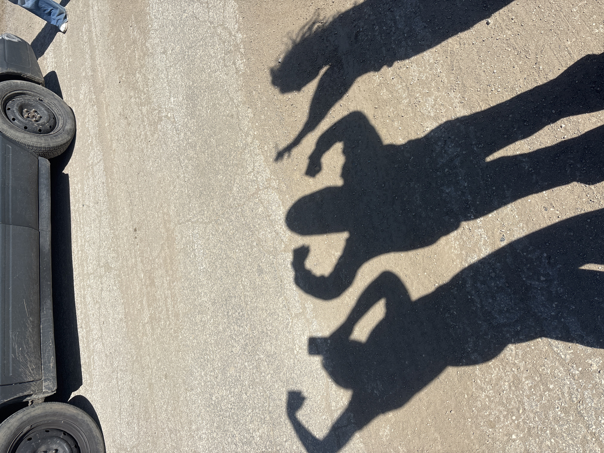 The image shows a section of asphalt street, with a portion of a dark-colored vehicle (wheel and body) on the left. There are shadows of three people standing near each other cast on the asphalt. A foot clad in a jean-covered leg and white shoe can be seen to the top left of the image. The location of Zhivkovo, Bulgaria is not verifiable using only the image provided.