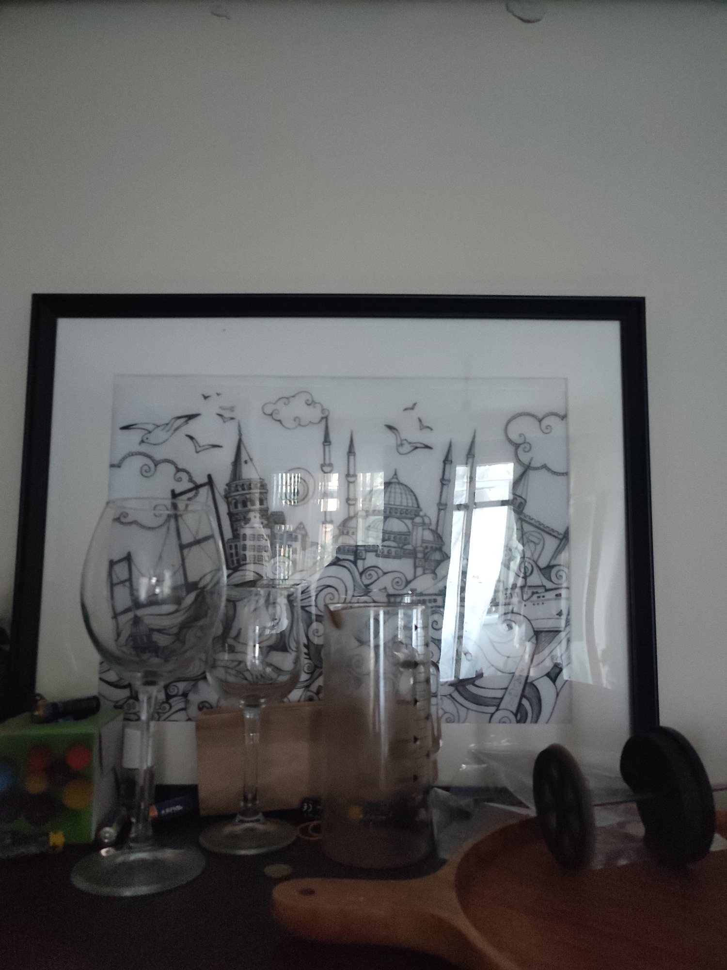 The image displays a framed black and white sketch of Istanbul's skyline, featuring prominent landmarks such as the Galata Tower and mosques. The artwork is positioned on a surface, likely a shelf or table, in front of a white wall. In the foreground, several objects are visible, including two wine glasses, one larger than the other, placed to the left of the artwork. To the right of the glasses, a cylindrical object, possibly a vase or a container, is present. The overall scene is dimly lit, with reflections on the glass of the artwork and the wine glasses suggesting ambient light sources. There are no people visible in the image. The presence of a sketch depicting Istanbul landmarks suggests a possible connection to the city. However, there is no information within the image itself that definitively places the scene in Kadıköy, Turkey.
