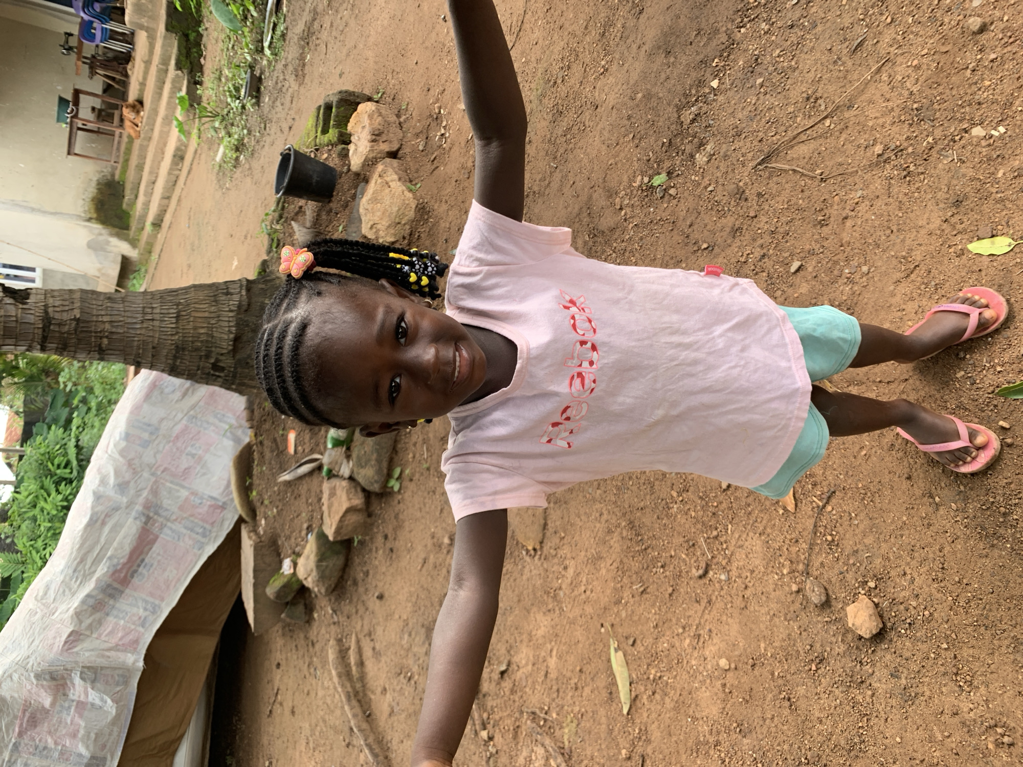A young Black girl with braided hair adorned with a butterfly clip stands in the foreground, facing the camera with her arms extended outwards. She is wearing a pale pink t-shirt with the word "Reebok" in red lettering and light blue shorts. Her feet are clad in pink flip-flops. She appears to be smiling. The ground beneath her is composed of reddish-brown dirt with scattered rocks and fallen leaves.
Behind the girl, to her right, a coconut palm tree trunk rises. Further back, a light-colored building with a white-framed window is visible, with a set of concrete stairs leading up to it. On the stairs, a brown dog lies down. To the left of the stairs, two wooden tables are present, one with a green bucket on it and the other with a sewing machine. In the middle ground, to the left of the girl, is a large, white, tarp-covered structure, possibly sheltering something. Dense green foliage can be seen in the background, particularly behind the tarp. The overall environment suggests an outdoor setting in Akwanga, Nigeria.