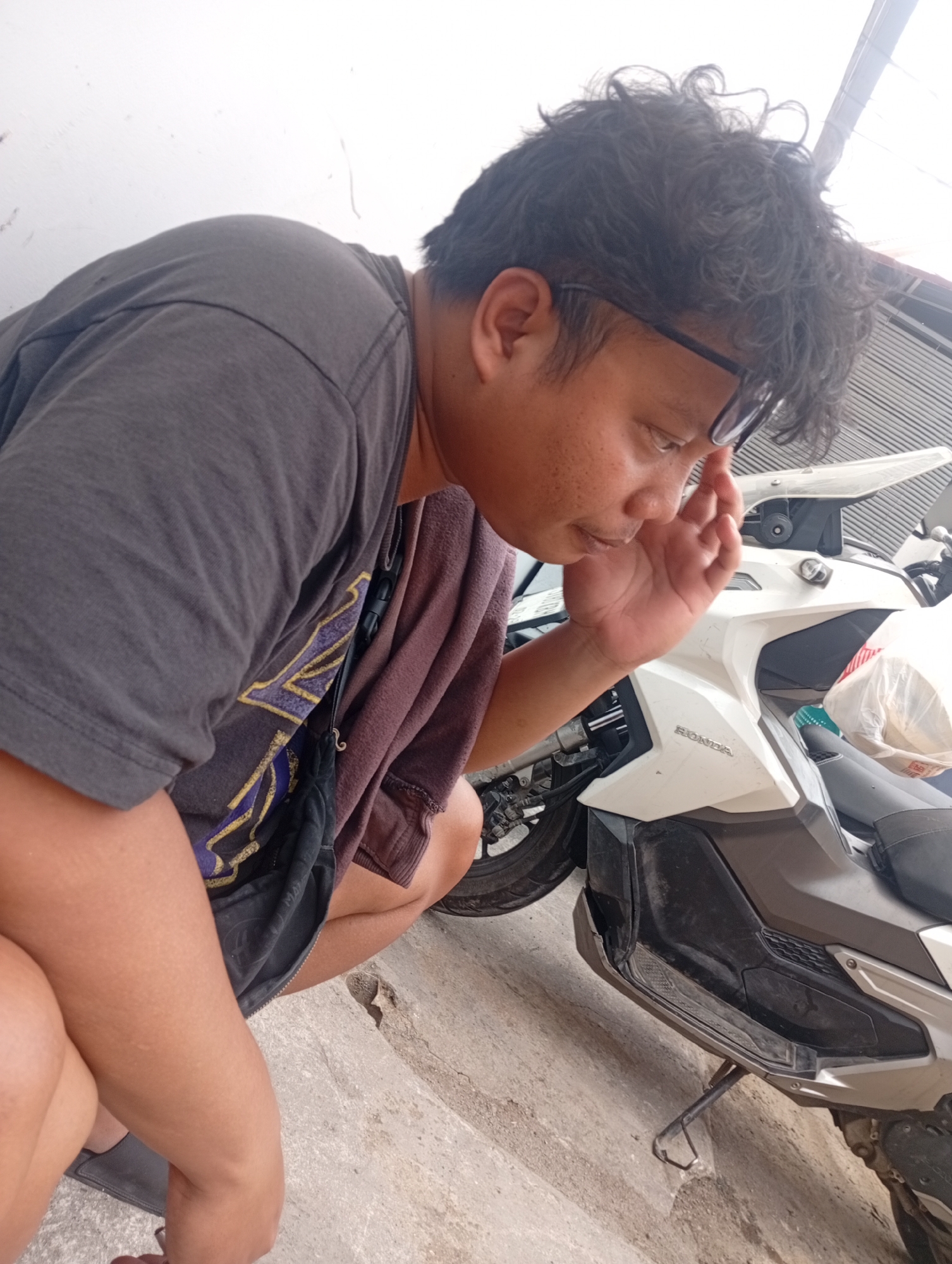 A person with curly dark hair and glasses is seen from the chest up, crouching beside a white motorcycle. The person is wearing a gray t-shirt with a yellow and purple design and a brown hooded jacket draped over their shoulders. They are looking down and touching their forehead with their right hand. The motorcycle has the "HONDA" logo visible on its side. The background shows a light-colored wall and a corrugated metal roof. The scene appears to be outdoors, possibly in a parking area or yard. The overall impression is of a casual, candid moment captured in an urban environment in Bekasi, Indonesia.
