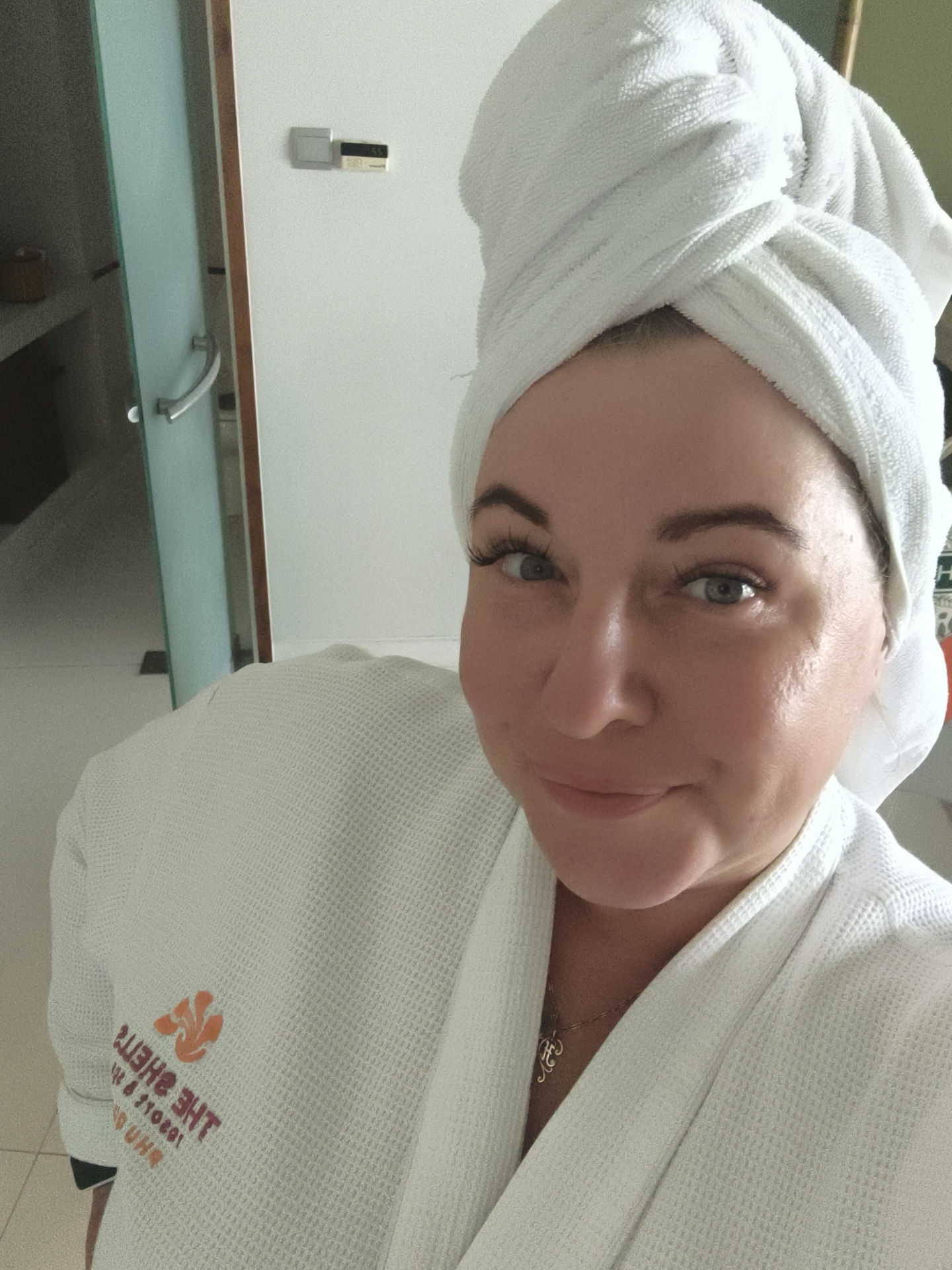 A woman is taking a selfie in what appears to be a hotel bathroom. She is wearing a white bathrobe and a white towel is wrapped around her head. The background shows a glass-enclosed shower area and a thermostat on the wall. The bathrobe has an embroidered logo which is partially visible and reads "THE SHELL." The woman is smiling and looking at the camera. The location is identified as Phú Quốc, Vietnam.