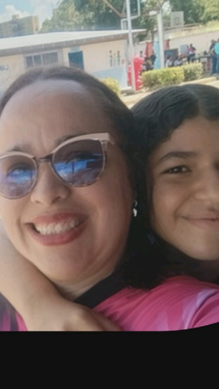 A close-up, outdoor, mid-shot photograph shows two females. The female on the left is wearing large, light-colored sunglasses with a tortoiseshell pattern and a bright pink and black collared shirt. She has dark brown hair and is smiling with her teeth showing. The female on the right has dark brown, curly hair and is also smiling. Both females are positioned close to each other, with their faces angled toward the camera. In the background, a low-rise building with a light-colored roof and a light-colored wall is visible. Partially obscured by the building are several people and some red structures. The environment appears to be a public space, possibly a schoolyard or park, with some green foliage on the right side of the image. The lighting suggests it is daytime. The photograph is a selfie or taken by someone standing nearby, capturing a moment of shared enjoyment between the two individuals.