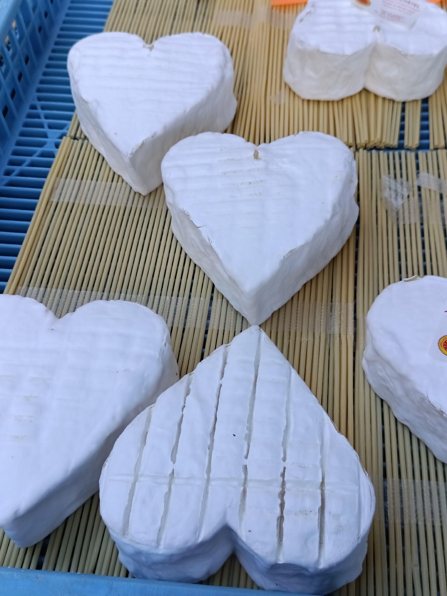 The image shows a display of heart-shaped cheeses on a mat made of thin wooden sticks. The cheeses are white with a soft rind. Some of the cheeses have crisscross scoring marks on their surface. The background includes a blue plastic crate. The scene appears to be a market stall. The location is identified as Le Neubourg, France, suggesting this is likely a local market or cheese shop. There are no people or actions depicted in the image.