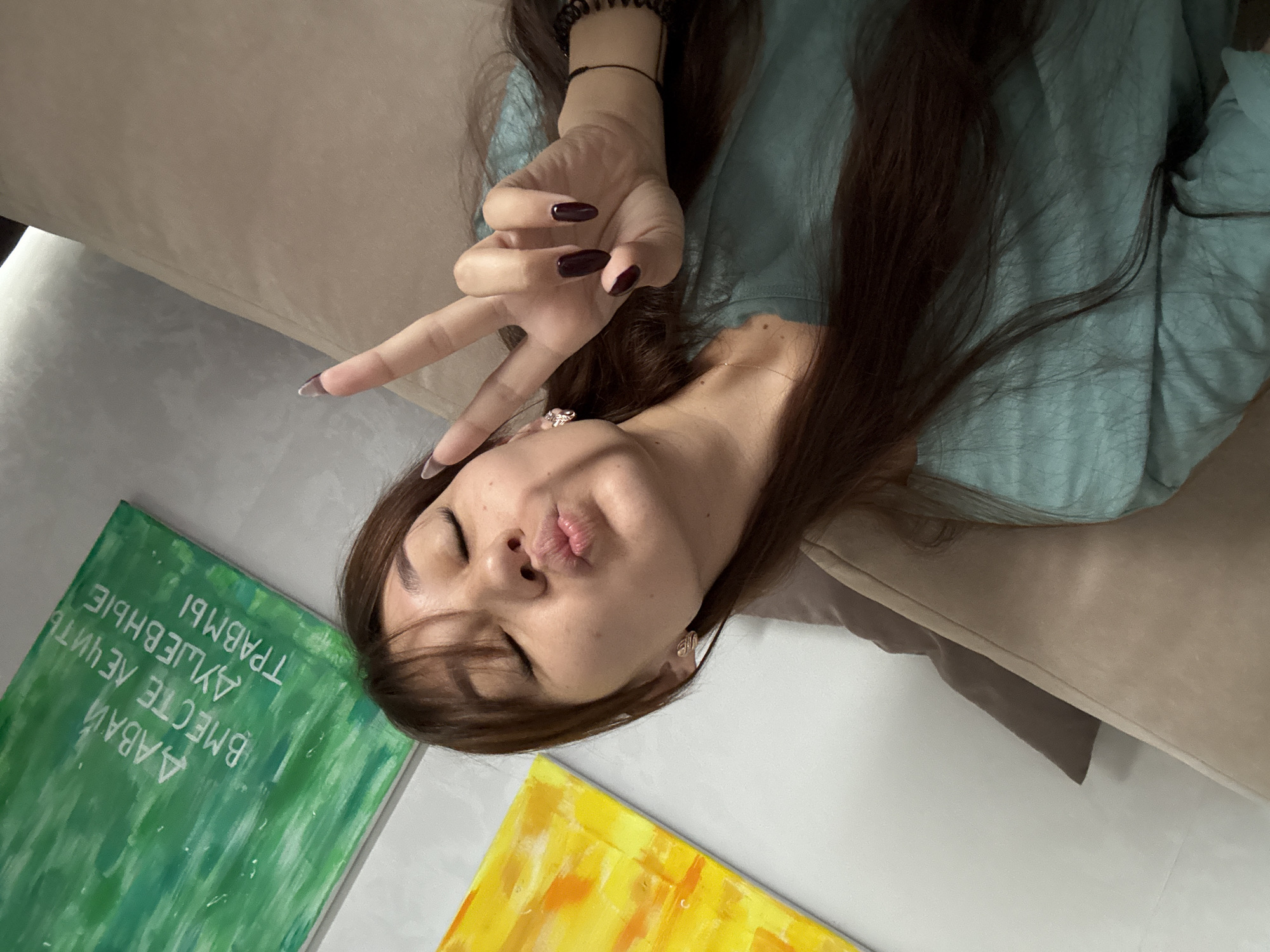 A woman with long, dark brown hair and bangs is lying on a couch, making a kissy face with her eyes closed and holding up a peace sign with her right hand. She is wearing a teal-colored t-shirt. Her fingernails are painted a dark maroon color. A thin necklace is visible around her neck. She has on small, sparkly earrings. Behind her, on the wall, are two paintings. The painting on the left is abstract with bright yellow and orange colors. The painting on the right is green with white text written in what appears to be Russian. The couch she is lying on is a light beige color. The overall scene appears to be an indoor residential setting, likely a living room. There is no indication of the location being specifically in the city of Novokuznetsk, Russia, as no landmarks or identifying features are visible that would confirm this.