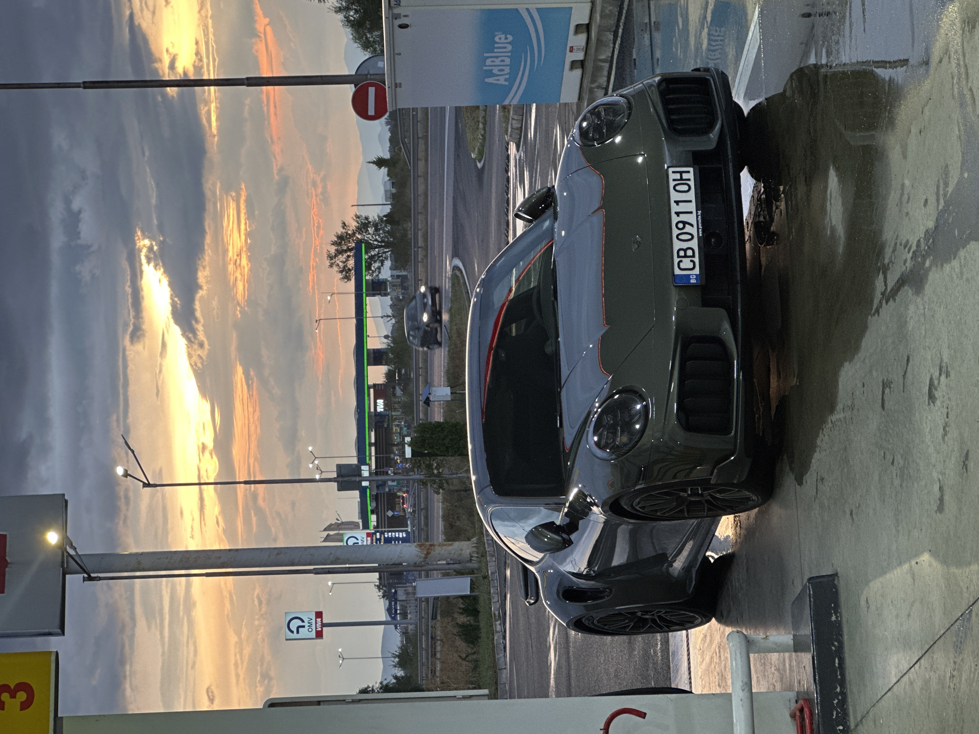 A dark grey Porsche 911 Turbo S is parked at a gas station in Drazhevo, Bulgaria. The car is positioned on a wet concrete surface, reflecting the surrounding lights and the vibrant sunset sky. The sky is filled with dramatic clouds illuminated by the setting sun, casting warm orange and yellow hues.
In the background, a highway overpass is visible with passing cars. A gas station sign with "OMV" branding is present, as well as an "AdBlue" dispensing unit. The overall environment suggests an evening scene at a roadside service area. The car's license plate reads "CB 0911 OH". There are no people visible in the image. The wet ground indicates recent rain or cleaning. The lighting suggests it is either dusk or dawn.