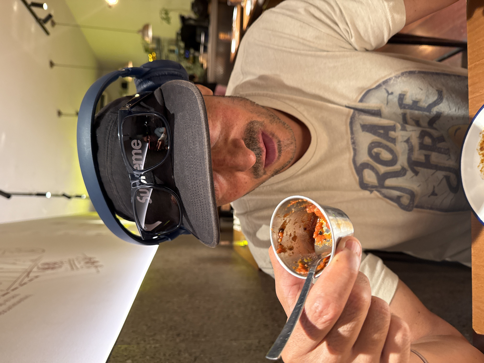 A man wearing a gray baseball cap, blue headphones, and sunglasses is holding a small metal container with food inside, and a spoon. He is wearing a light beige t-shirt with a graphic design. The location is Barcelona, Spain.