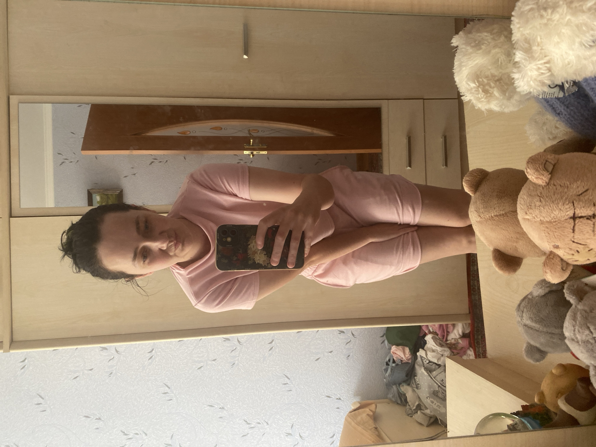 In a dimly lit bedroom, a young woman with her dark hair pulled up in a messy bun stands in front of a tall, wooden wardrobe with a mirrored door, taking a selfie with her phone. She is wearing a pale pink t-shirt and matching shorts. Her expression is one of slight concern. The wardrobe's mirror reflects the room behind her, revealing a floral patterned wallpaper and a closed wooden door with decorative glass inlays. To the left of the wardrobe, a messy pile of clothes is visible, and below it, a wooden shelf unit. In the foreground, reflected in the mirrored wardrobe door, are several plush teddy bears of various sizes and colors. One bear is predominantly beige, another is gray, and a white bear is visible to the right. The overall impression is a candid, everyday moment captured in a personal space.