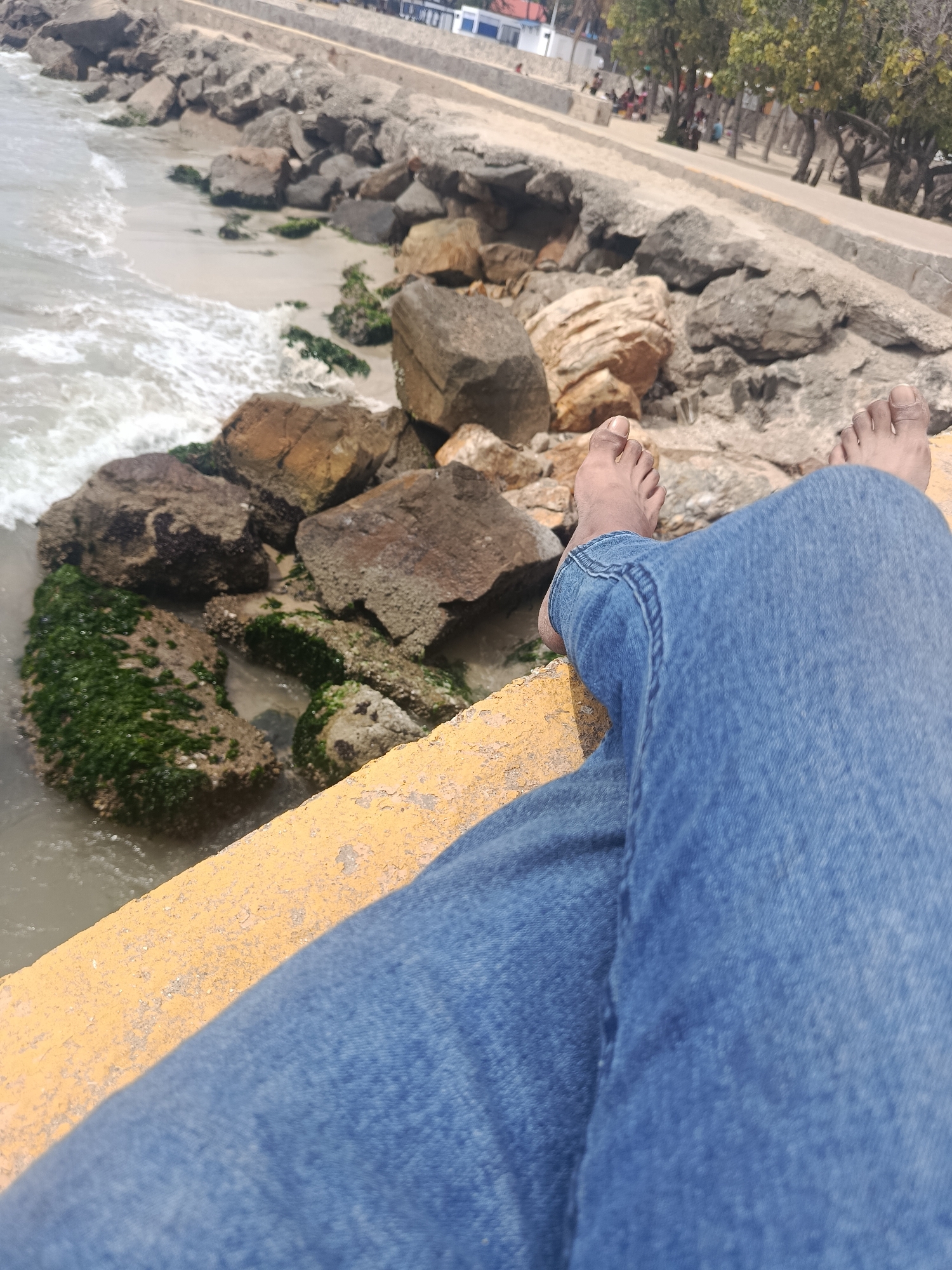 A first-person perspective photograph shows a pair of legs clad in blue jeans extending into the frame, with bare feet resting at the edge of a concrete barrier. The barrier has a visible yellow painted surface. Beyond the barrier, a coastal scene unfolds, including a beach with rocks and incoming waves. Patches of green algae are visible on some rocks. The background features a paved walkway with trees and some indistinct figures. The location is Juan Griego, Venezuela, as indicated by the coastal environment and architectural details in the background.