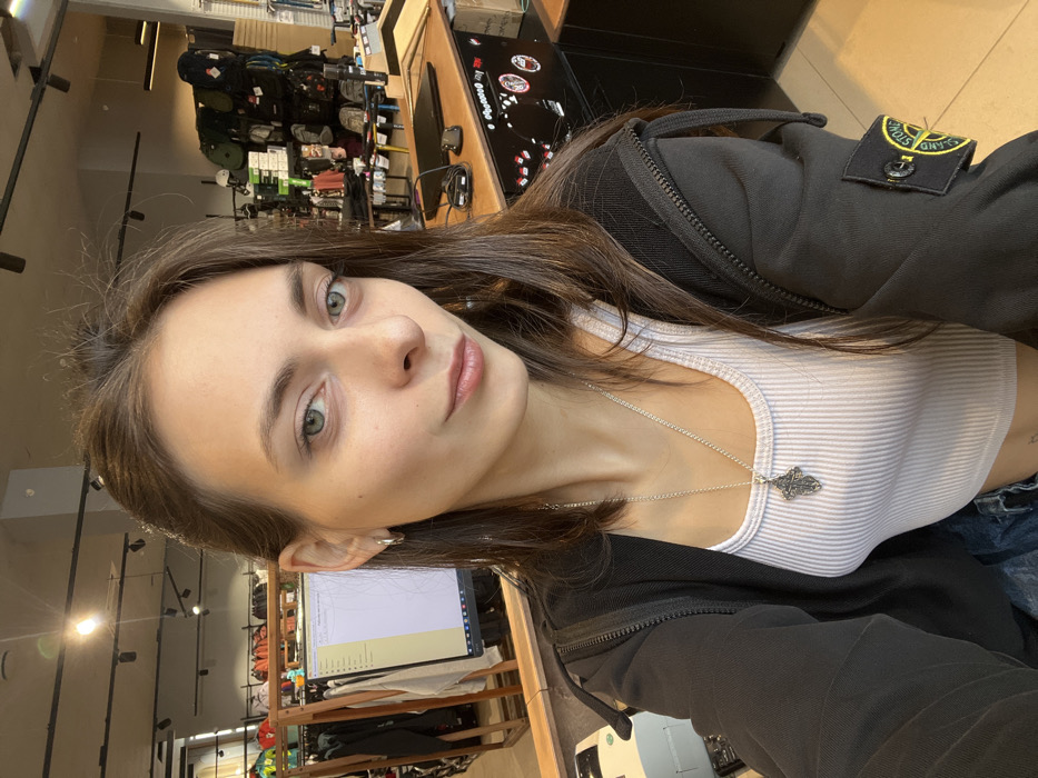 A young woman poses for a selfie indoors, partially turned. She wears a white ribbed tank top, a black jacket with a logo patch on the shoulder, and a silver necklace with an ornate pendant. Her hair is brown, and she has green eyes. The background shows a store interior with shelves of merchandise, a counter, and a computer monitor.