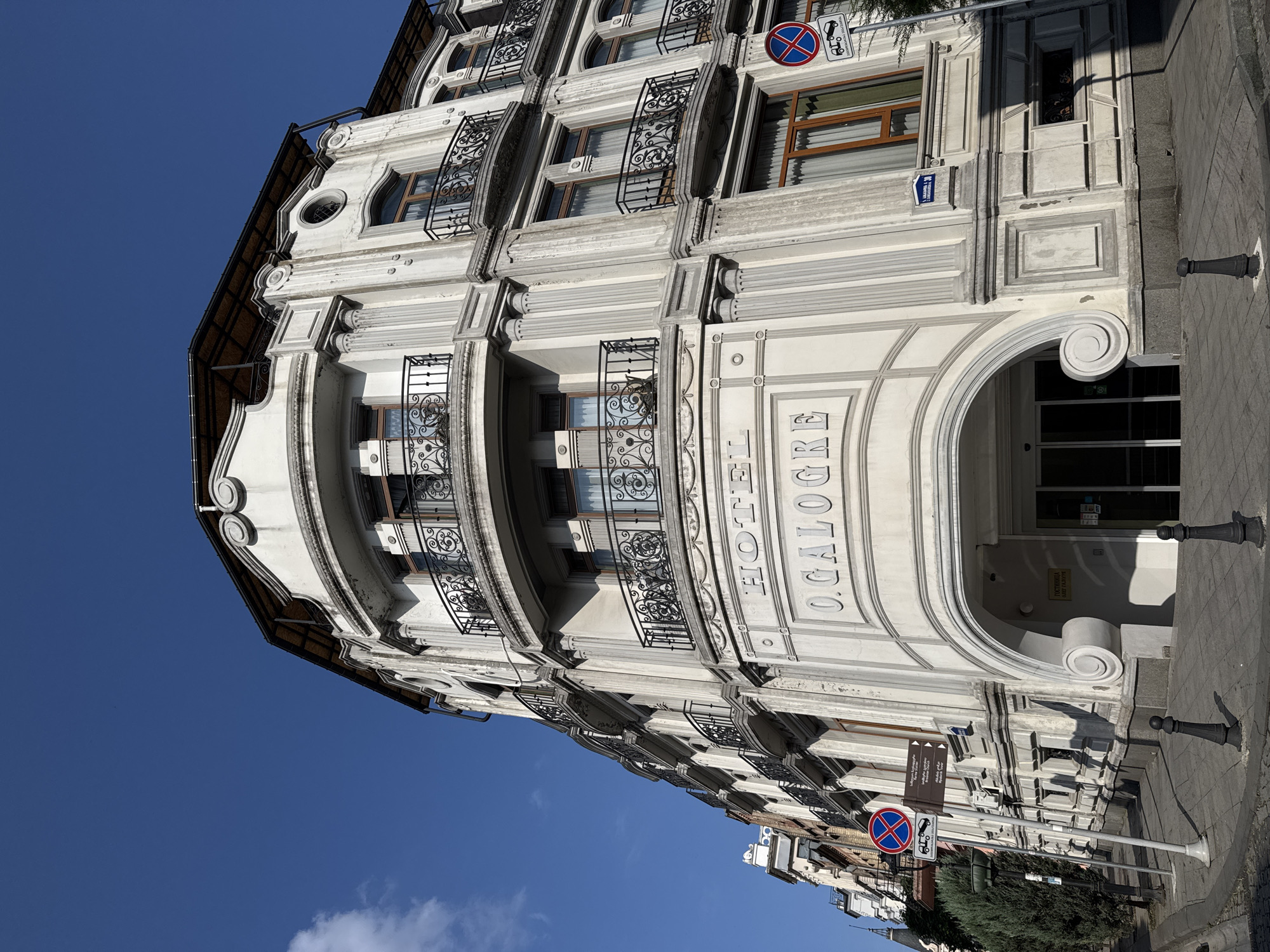 The image shows a white, ornate hotel building with multiple stories and balconies featuring wrought-iron railings. The hotel's name, "HOTEL O.GALGRE," is inscribed in relief above the entrance. The architecture suggests an older, European style. Street signs are visible, including one with Georgian script, indicating a location in Georgia. A blue street sign with the number "30" is mounted on the building. The building is situated on a city street with a sidewalk, a street lamp, and decorative bollards. The sky is clear and blue with a few wispy clouds. The overall scene depicts a well-maintained urban environment on a sunny day. This is Hotel O.Galgre located in Batumi, Georgia.