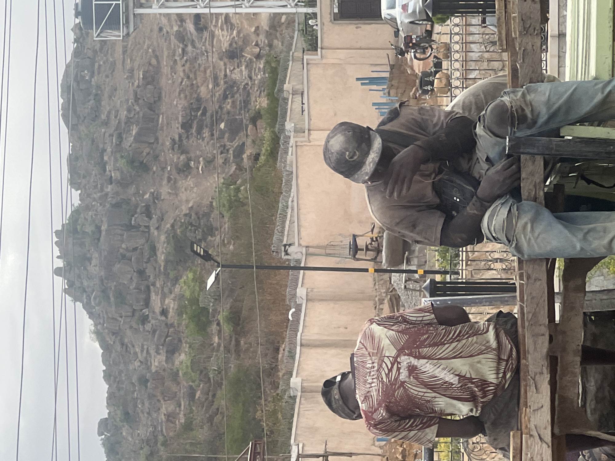 The image, rotated 90 degrees counter-clockwise from its natural orientation, depicts an outdoor scene with two men resting in the foreground against a rocky mountainous backdrop. When viewed upright, the man on the right is seen from behind, wearing a light-colored, short-sleeved shirt with a reddish-brown leafy print and a dark cap or helmet. He is seated on a rough wooden structure.
To his left, another man is also seated on the same wooden structure, facing partially away from the camera. He wears a light-brown long-sleeved shirt, distressed blue jeans with visible rips, and a dark cap. A dark shoulder bag is slung across his body. He is leaning forward with his hands resting, suggesting a moment of rest. A third person, wearing light-colored ripped jeans, is partially visible behind him.
In the middle ground, behind the men, there is a light-colored concrete building with vertical blue painted accents. A concrete wall, topped with barbed or razor wire, runs in front of the building. A tall black streetlight with a rectangular white panel stands prominently.
The background is dominated by a large, rugged, dark gray rocky mountain or hill under an overcast sky. Power lines or cables stretch across the upper portion of the scene. Parts of a white vehicle and a motorcycle are visible parked near the building on the left. The scene suggests a daytime setting in a semi-urban or rural area, typical of landscapes in Jos, Nigeria, where the men might be taking a break from work. There is no visible text.