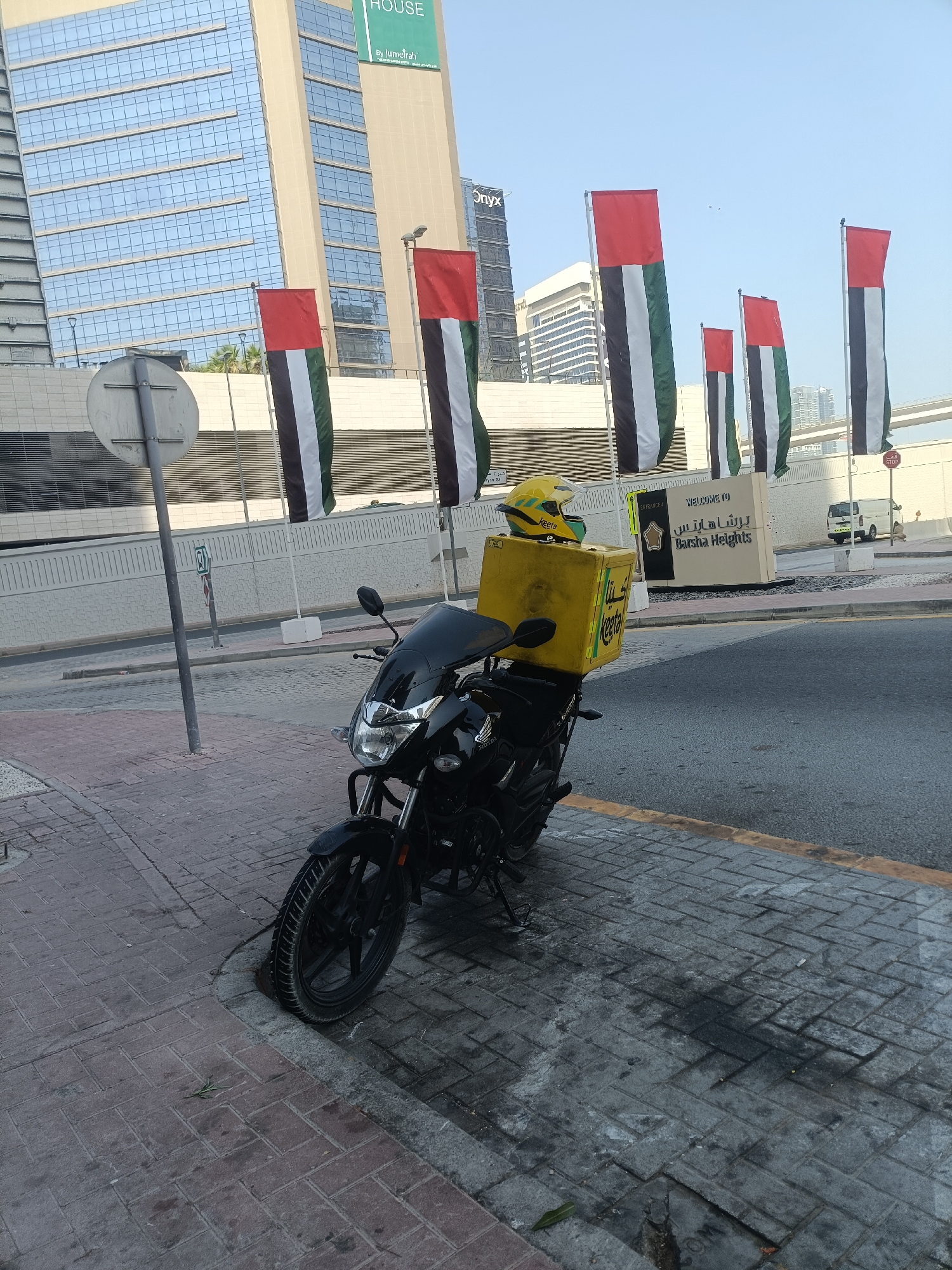 A black motorcycle is parked on a paved sidewalk next to a street. The motorcycle has a yellow delivery box mounted on the back and a yellow helmet resting on top of it. In the background, several large flags with the colors red, white, green, and black are displayed vertically. Behind the flags, there are modern buildings, including one with a sign that reads "Barsha Heights." A white van is visible further down the street. The scene appears to be an urban environment in Dubai, United Arab Emirates, likely in the Barsha Heights area given the signage.