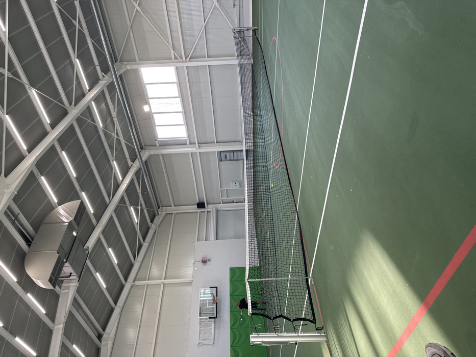An interior view of a modern indoor sports facility in Gorna Mitropolia, Bulgaria, presents a tennis court with a green hard surface marked with white lines for tennis and additional red lines. A black tennis net is stretched across the court, secured by silver posts. One yellow tennis ball rests on the court beyond the net, while another is positioned mid-air near an adult male on the far side of the court. This individual, dressed in dark clothing, is partially obscured but appears to be holding a tennis racket, positioned as if to strike the ball.
The facility features high, gabled ceilings with exposed white metal trusses supporting numerous linear fluorescent lights and a large, curved silver duct system. The walls are constructed from white corrugated metal panels. On the left wall, a basketball hoop with a clear backboard and orange rim is mounted above a green padded wall section. A large grey roll-up door and a standard white door are visible on the far wall. A series of rectangular windows situated high on the right wall admit natural light. The floor shows subtle areas of discoloration and wear. The toe of a grey shoe is visible in the bottom left foreground, indicating the photographer's close proximity to the court.