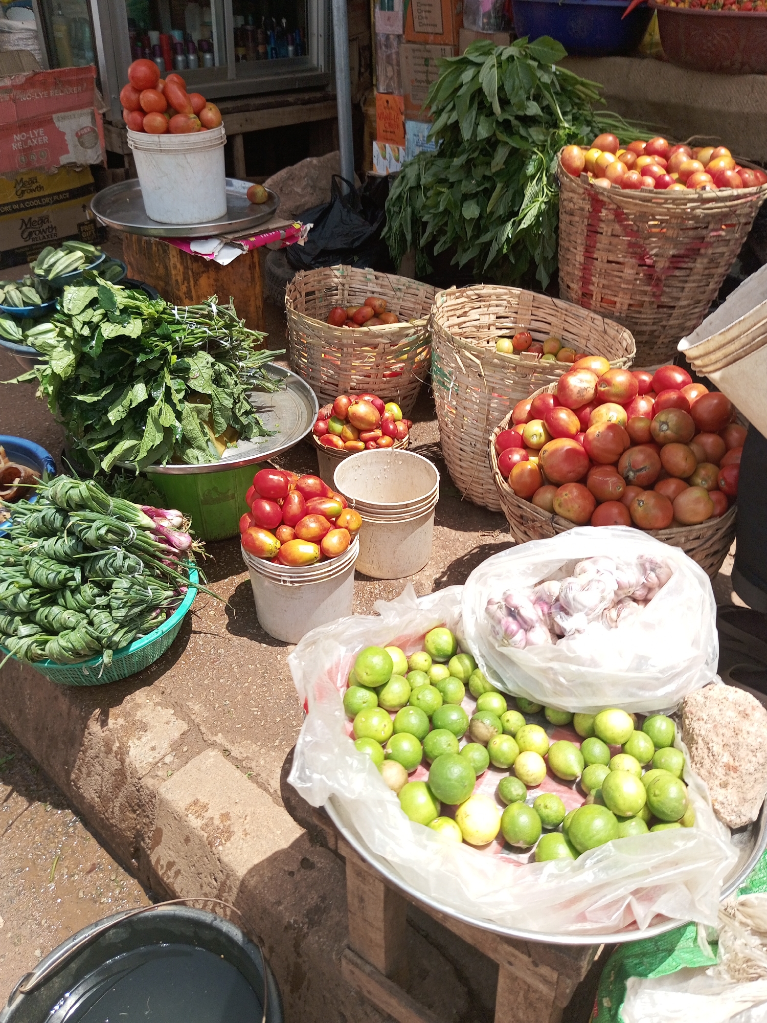 This is a medium-shot, eye-level, outdoor photograph of a market stall in Bukuru, Nigeria. The scene is filled with a variety of fresh produce, suggesting it is a vibrant, bustling marketplace. The sun is shining, casting distinct shadows, indicating it is likely daytime.
Dominating the foreground are several large baskets and buckets overflowing with fruits and vegetables. To the left, a green plastic basket holds bundles of leafy greens, possibly spinach or amaranth, tied together. Next to it, a white bucket is filled to the brim with bright red tomatoes. Further to the right, a large woven basket is packed with more tomatoes, varying in shades of red and orange. In front of this, another woven basket contains a mix of smaller tomatoes and what appear to be limes or small green citrus fruits. A large white plastic bag is also filled with these green fruits, and a few more are scattered on a metal tray. Another white bucket sits empty. Behind the tomatoes, a large pile of green leafy vegetables is visible. In the lower right corner, a white plastic bag contains several heads of garlic, and a light-colored rock is also present. To the far left, a dark bucket holds some liquid.
In the background, a market stall structure is visible, with shelves holding various packaged goods. Some boxes with text, including "NO-LYE RELAXER" and "Mega Growth," are stacked against the stall. The overall impression is one of abundance and freshness, characteristic of a local food market. The scene is well-lit, with natural daylight illuminating the produce. No people are visible in the frame.