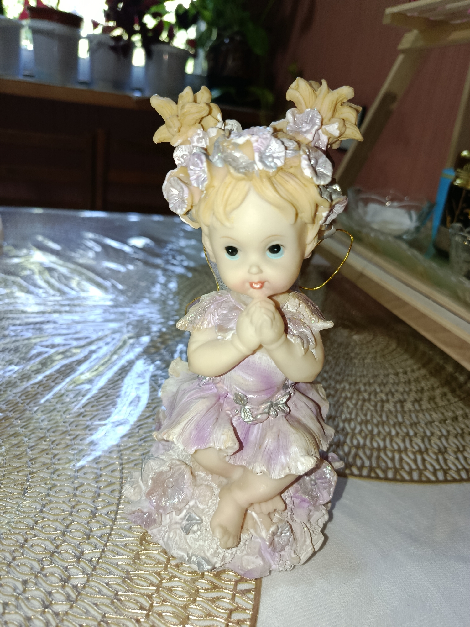 A small, porcelain figurine of a cherubic fairy with blonde hair styled in twin buns adorned with flowers and silver accents sits on a textured, golden placemat. The fairy wears a pale lavender, petal-shaped dress and has its hands clasped in front of its chest in a praying or hopeful gesture. Its eyes are a bright blue, and it has a slight smile. The background includes a wooden shelf with potted plants, a glimpse of a room with wooden furniture, and a reflective surface on the table. The scene appears to be indoors, possibly in a home environment. There is no indication of location in Tashkent, Uzbekistan.