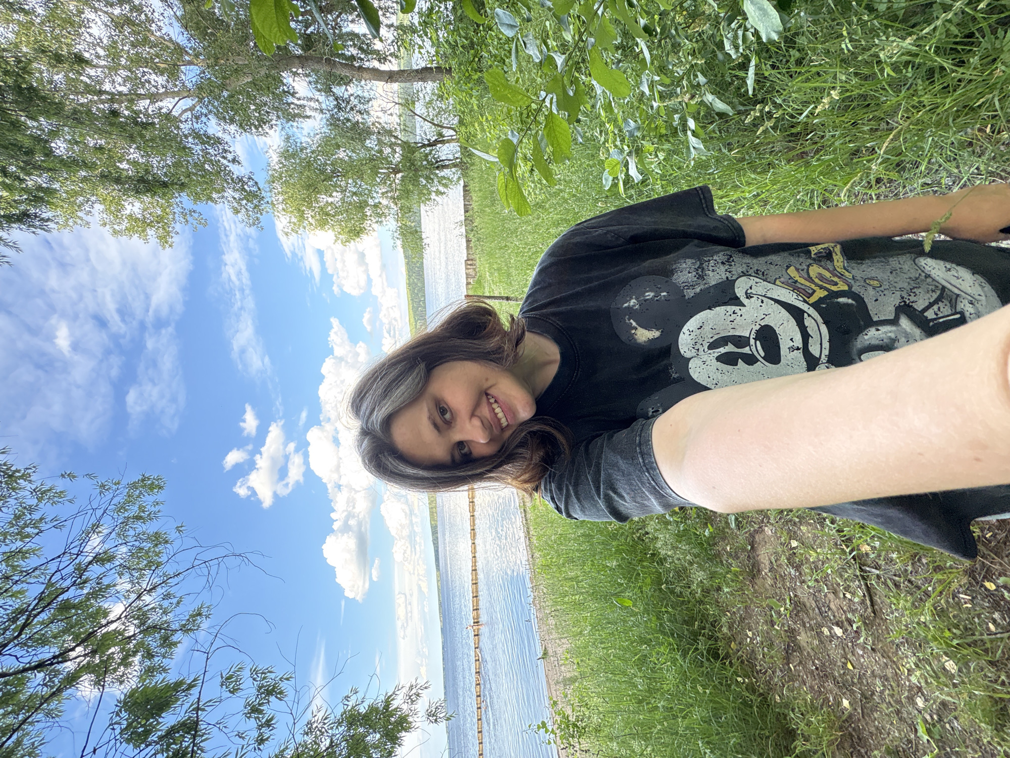 In the city of Chaikovsky, Russia, a woman with shoulder-length brown hair and subtle grey streaks smiles at the camera. She is wearing a black t-shirt with a graphic of Mickey Mouse. The woman is holding up her arm, as if taking a selfie, and her arm and a portion of the t-shirt are in the foreground.
The background suggests an outdoor scene, possibly a park or natural area near a body of water. Tall green grass and trees are visible, with a calm blue lake or river stretching out behind the woman. A wooden barrier or row of posts can be seen in the water, and there appear to be a few people in the distance, suggesting a recreational area. The sky is a bright blue with scattered white clouds, indicating fair weather. The overall impression is a casual, summery day by the water.