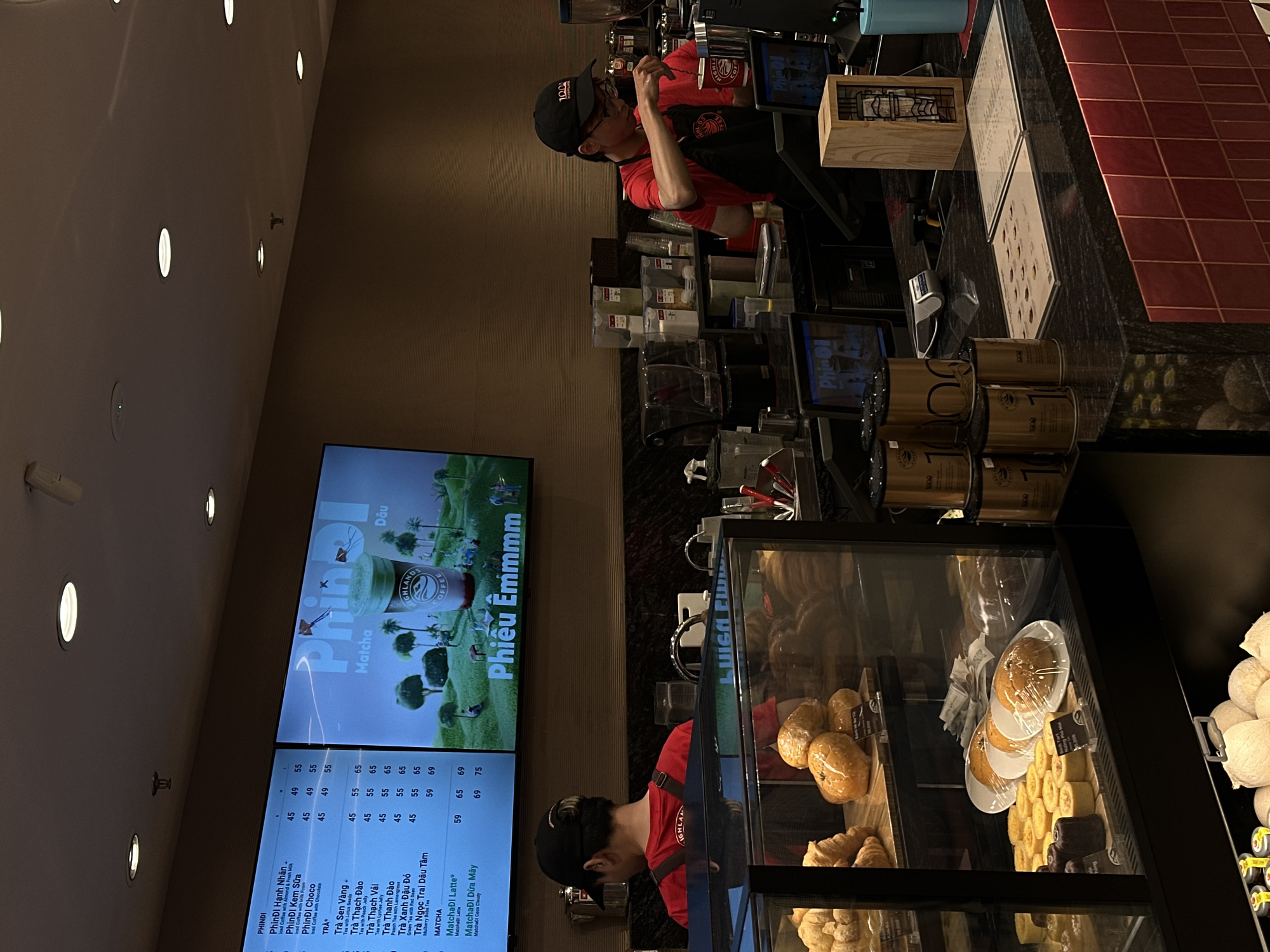 The image depicts the interior of a cafe in Da Nang, Vietnam. The scene is dominated by a counter area where two baristas, dressed in red t-shirts and black aprons, are working. One barista, wearing a black baseball cap, is actively preparing a drink. The other barista, also wearing a cap, is partially visible behind the counter, facing away from the camera.
The cafe setting features a large digital menu board displaying drink options, with the prominent words "PhinĐI" and "Phiêu Êmmmm" visible. Below the menu, a list of drink names in Vietnamese, such as "Trà Sen Vàng" and "MatchaÐI Latte*", along with prices, can be seen. In the foreground, a glass display case showcases an assortment of baked goods, including croissants, buns, and cake rolls, suggesting a bakery component to the cafe.
Behind the counter, shelves are stocked with various containers, cups, and coffee-making equipment. Several cylindrical canisters with "1000" printed on them are stacked on the counter. The overall lighting suggests it might be daytime, with artificial lights illuminating the interior. The atmosphere appears to be busy and functional, typical of a cafe during operating hours. The presence of Vietnamese text on the menu and the general ambiance strongly indicate a location within Vietnam.