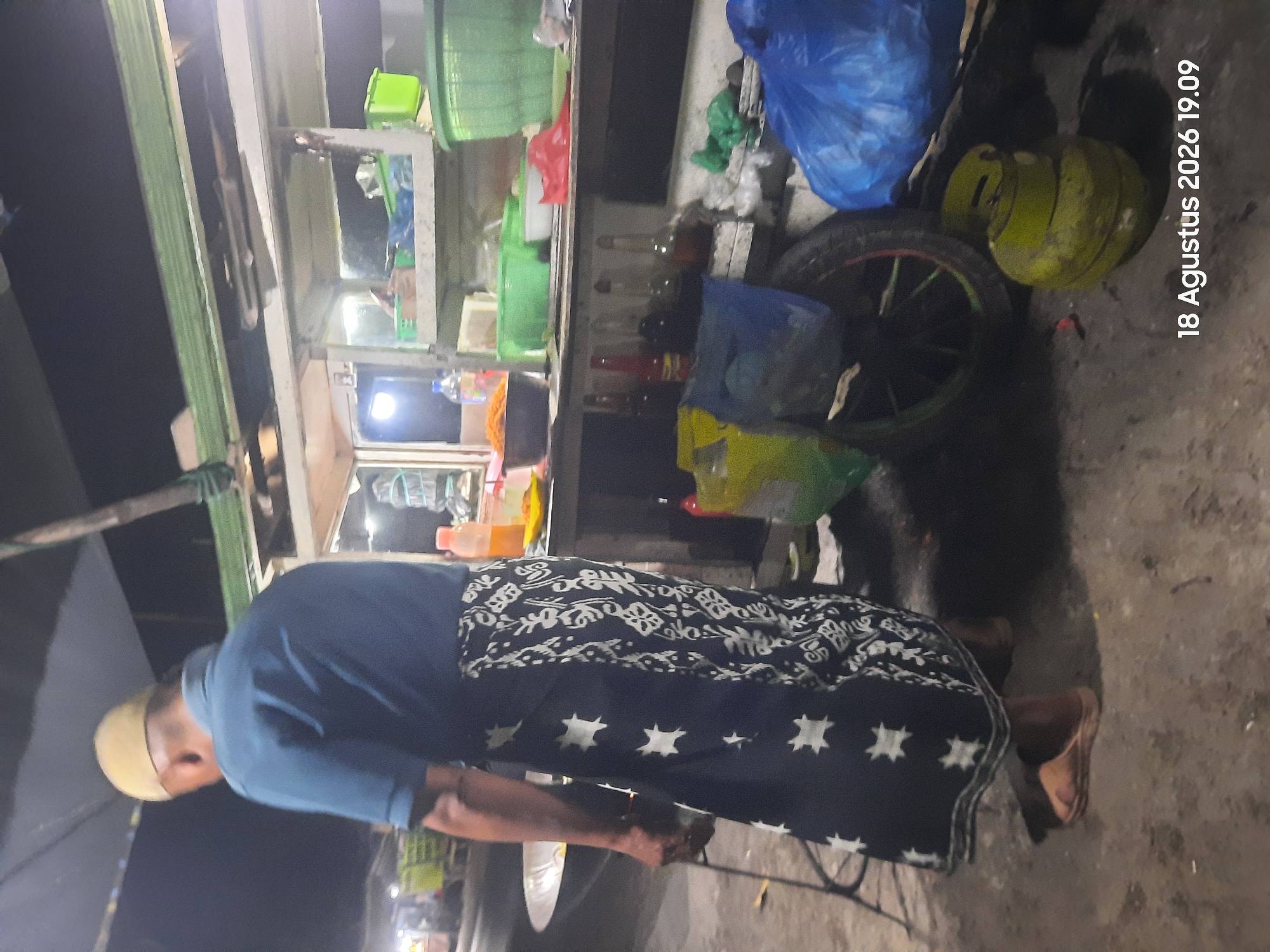 The image depicts an outdoor scene at night, likely in Sukokerto, Indonesia. A person wearing a blue shirt and a traditional sarong with a white star pattern stands in front of a food stall. The stall has shelves stocked with various items, including bottles, containers, and what appear to be food products. In the foreground, a green gas cylinder is visible on the ground, alongside a black tire and some plastic bags. The lighting suggests it's a well-lit street or market area. The date and time stamp "18 Agustus 2026 19.09" is superimposed on the bottom right of the image.