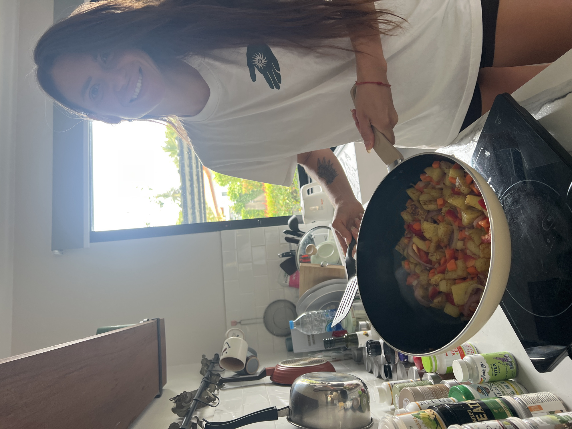 A woman with long brown hair is standing in a kitchen, smiling at the camera as she cooks. She is wearing a white t-shirt with a black graphic on it and black shorts. In her hands, she holds a spatula, stirring the contents of a frying pan filled with chopped vegetables, including potatoes, carrots, and red onions. The frying pan is on a black induction stovetop. To the left of the stovetop, a counter is lined with various supplement bottles. Behind the woman, a window offers a view of lush greenery and a building, suggesting a tropical location. The kitchen walls are tiled with white subway tiles, and various kitchen utensils and cookware are visible on a rack and shelves. The overall scene is bright and airy, with natural light streaming in from the window. The specific location is identified as Baan Bang Rak, Thailand.