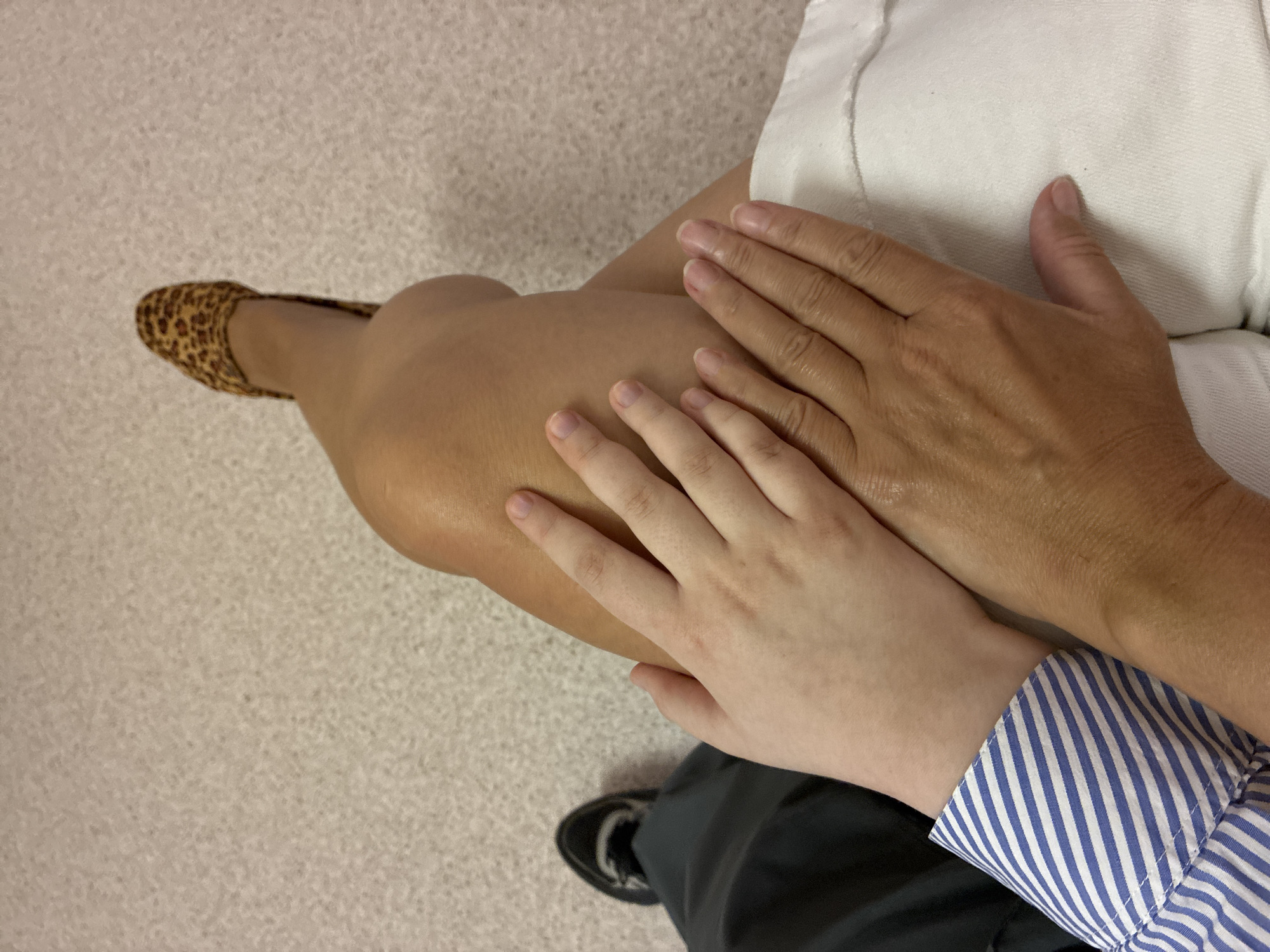 A close-up shot shows two pairs of hands resting on a person's lap. The upper hands, belonging to a child, are light-skinned with short fingernails. They rest on the lower hands, which are tanned and have manicured nails. The person's legs are visible, one with a leopard-print shoe. The background is a textured carpet.