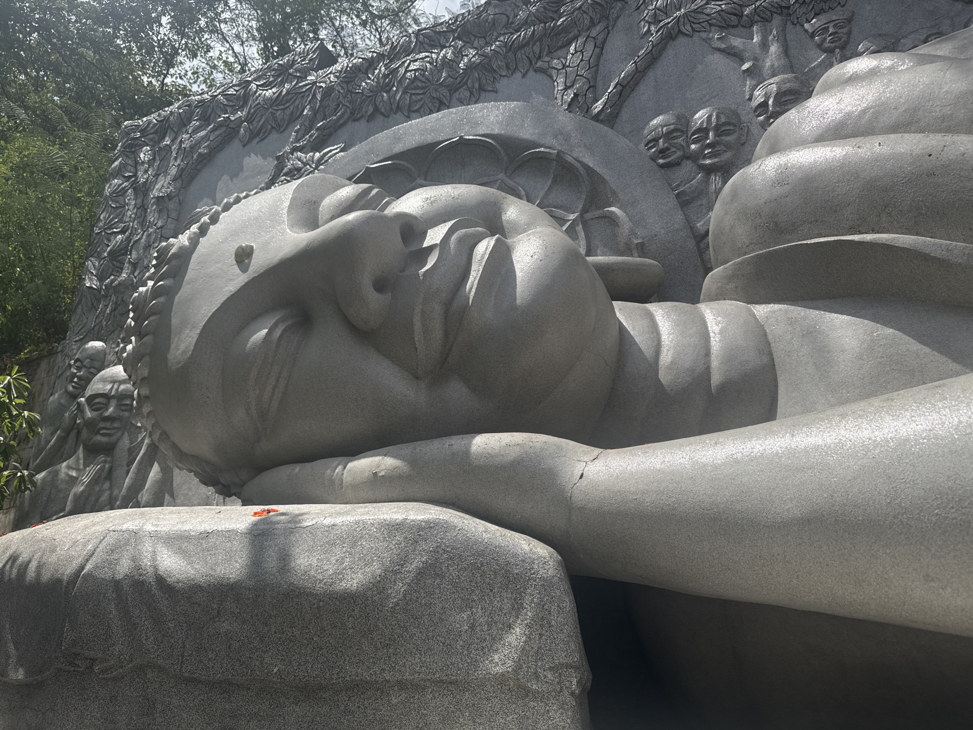 A large, gray, reclining Buddha statue is the main subject of this outdoor image, set against a background of lush green trees and a bright sky. The statue is carved from stone and depicts the Buddha in a peaceful repose, with his head resting on his hand. The detail of the carving is evident in the folds of his robes and the serene expression on his face. Behind the Buddha, a stone relief depicts several smaller figures, possibly disciples or celestial beings, looking on. The overall atmosphere is one of tranquility and reverence. The statue appears to be situated in a temple or a sacred garden, given the religious iconography and the natural surroundings.