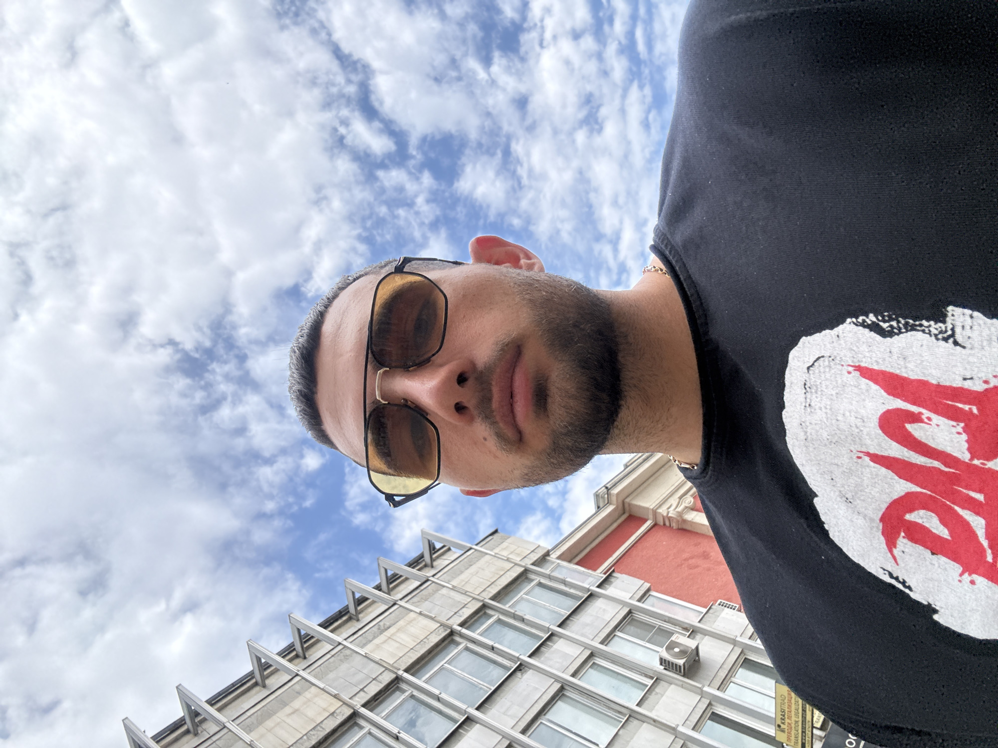 The photograph depicts a low-angle selfie of a man wearing sunglasses and a black t-shirt with a red and white graphic. He is facing downwards towards the camera. Behind him, the sky is visible with scattered white clouds against a blue background. A multi-story building can also be seen in the background. The building has a facade of gray stone with rows of windows and a section of red brick on its upper floors. Based on the architectural style and signage visible on the building, the location is likely in Ruse, Bulgaria. The building is located near the intersection of ul. "Knyaz Heski" and ul. "Tsar Kaloyan."